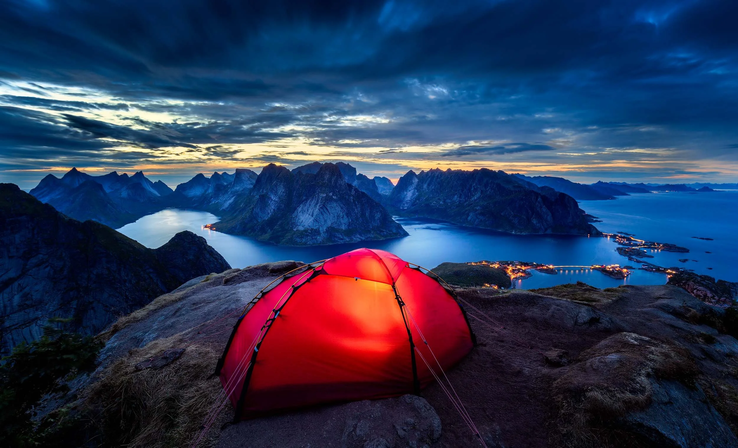 Illuminated red tent at the top of Reinebringen, Lofoten, Norway during sunset showcasing the spectacular surrounding landscape with steep mountains and fjords. 