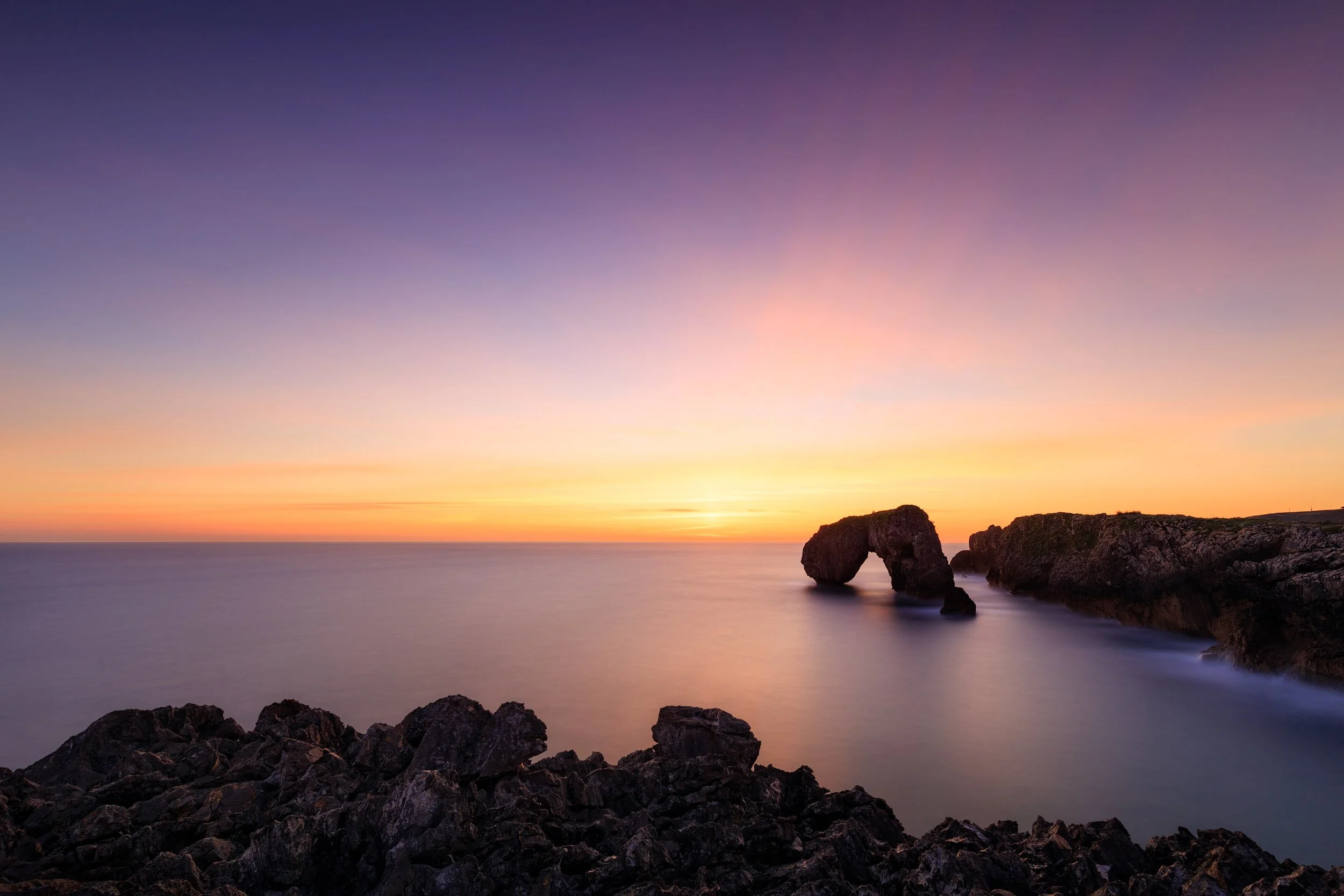 Long exposure of the beautiful rock arch of Castro de Las Gaviotas located near Punta de Huelga near the village of Villahormes in the autonomous community of Asturias in Spain during sunrise. The water of the atlantic ocean is blurred due to the lon