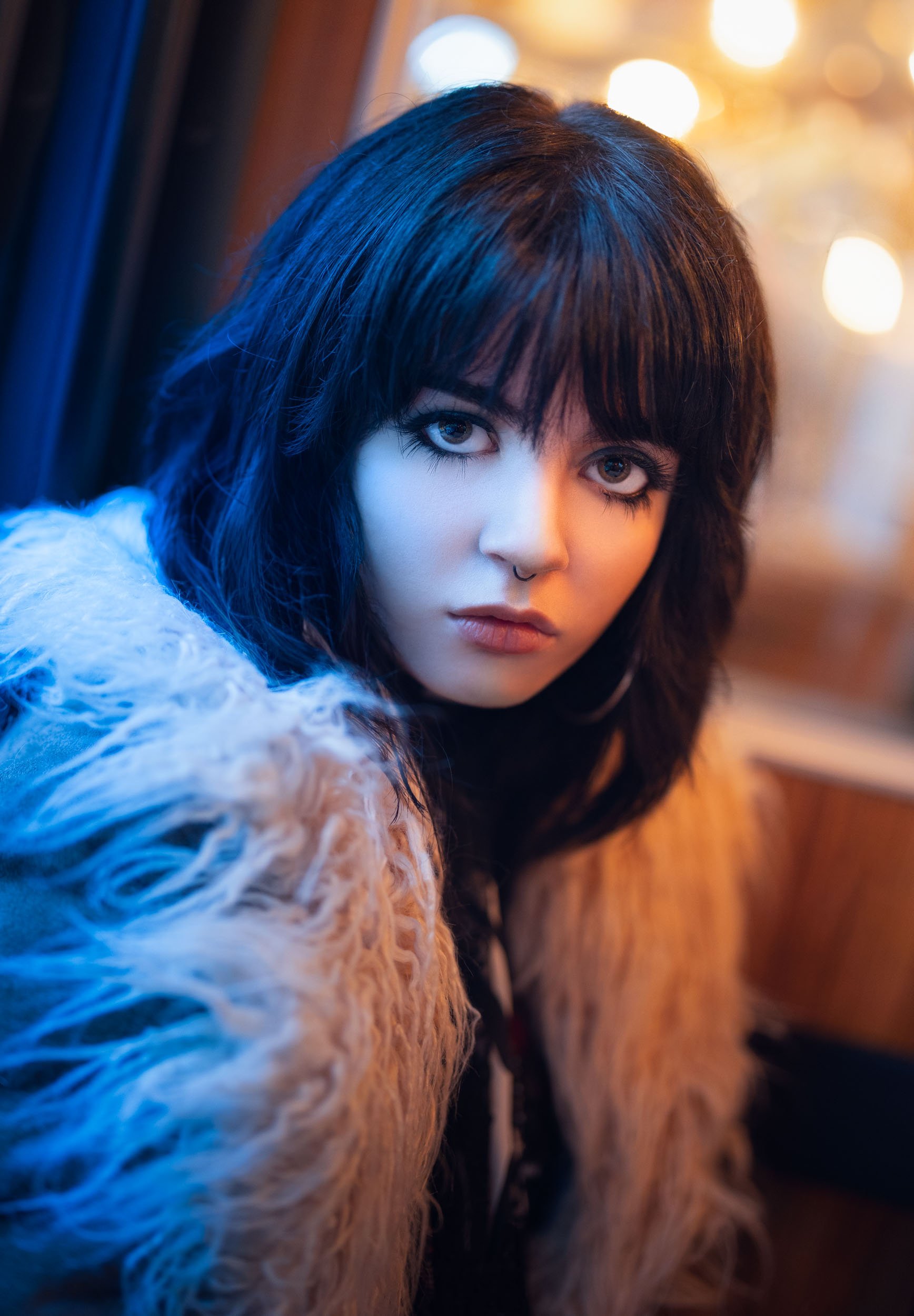 Indoor close-up portrait of a young, beautiful girl with large, sad eyes and shoulder-long black hair wearing a fahsionable fur-coat. Her face is partly illuminated by the cold light coming in from the window and the warm lights of the room. 