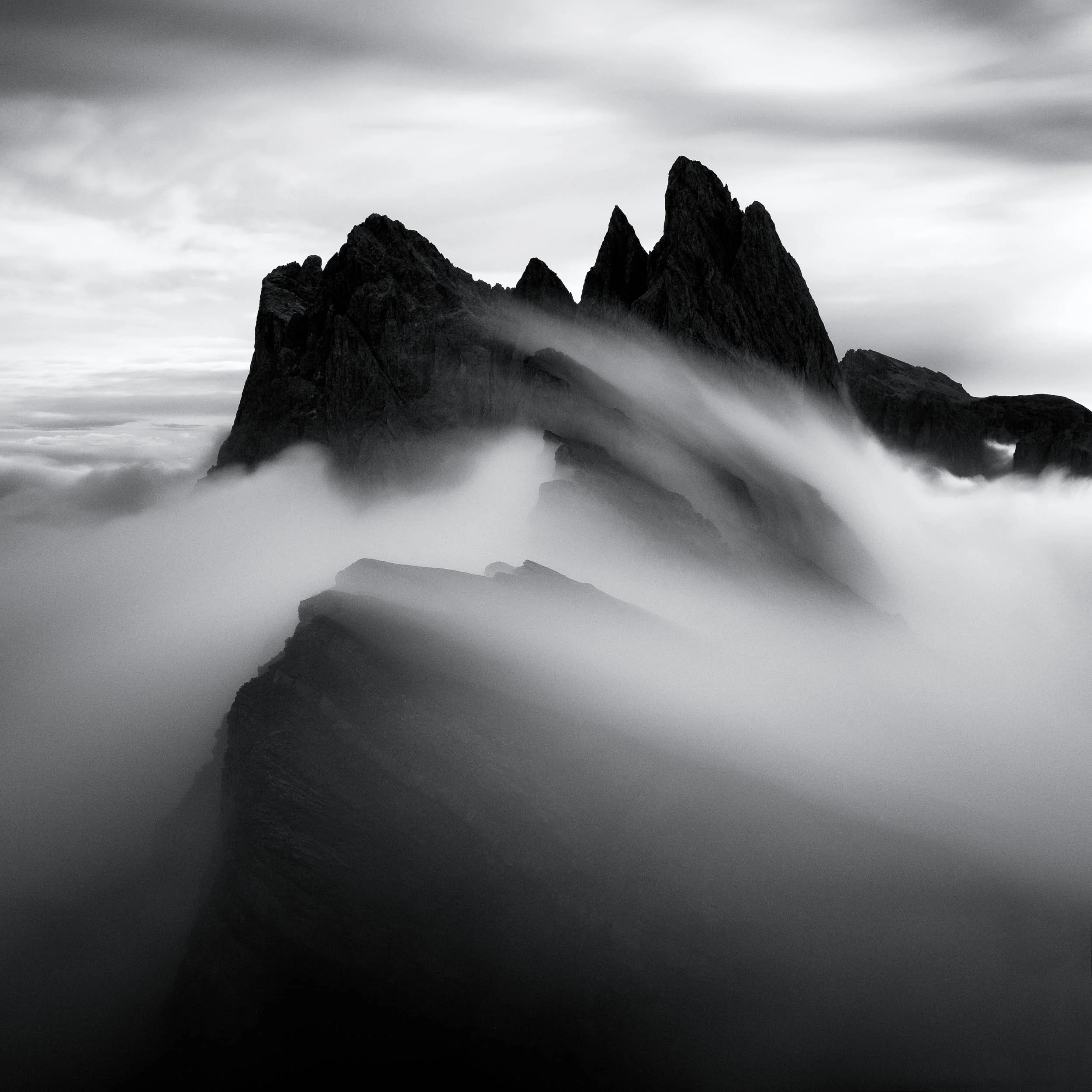 Black and white fine-art capture in square-crop of the Seceda ridgeline in the Dolomites, Italy. A stream of clouds is creeping up towards the peaks of the Geisler peaks. 