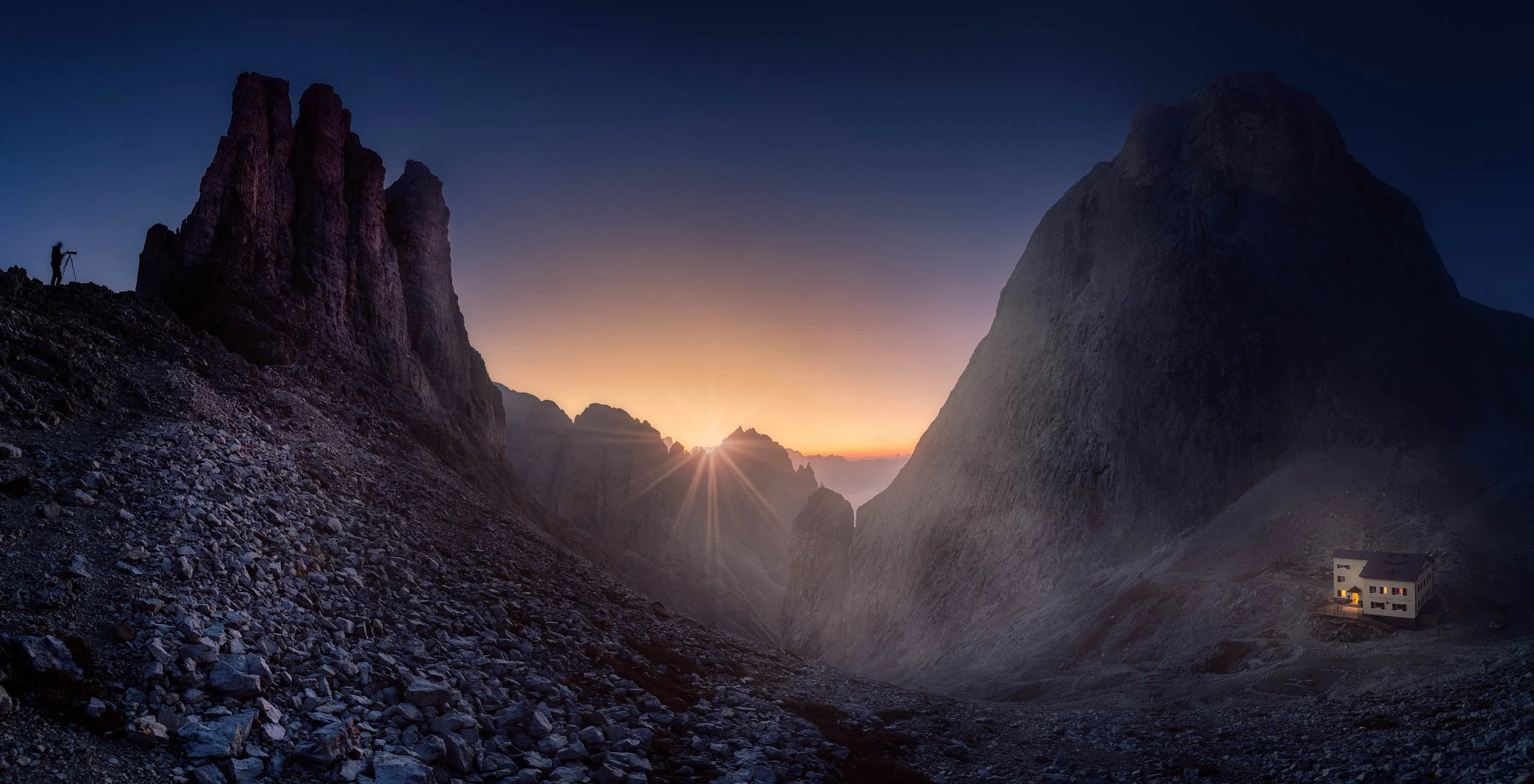 A photographer having his camera set-up on his tripod to take pictures of the sunrise at the Vajolet Towers and the mountain hut Rifugio Re Alberto in the Rosengarten mountain group in the Dolomites, Italy. 