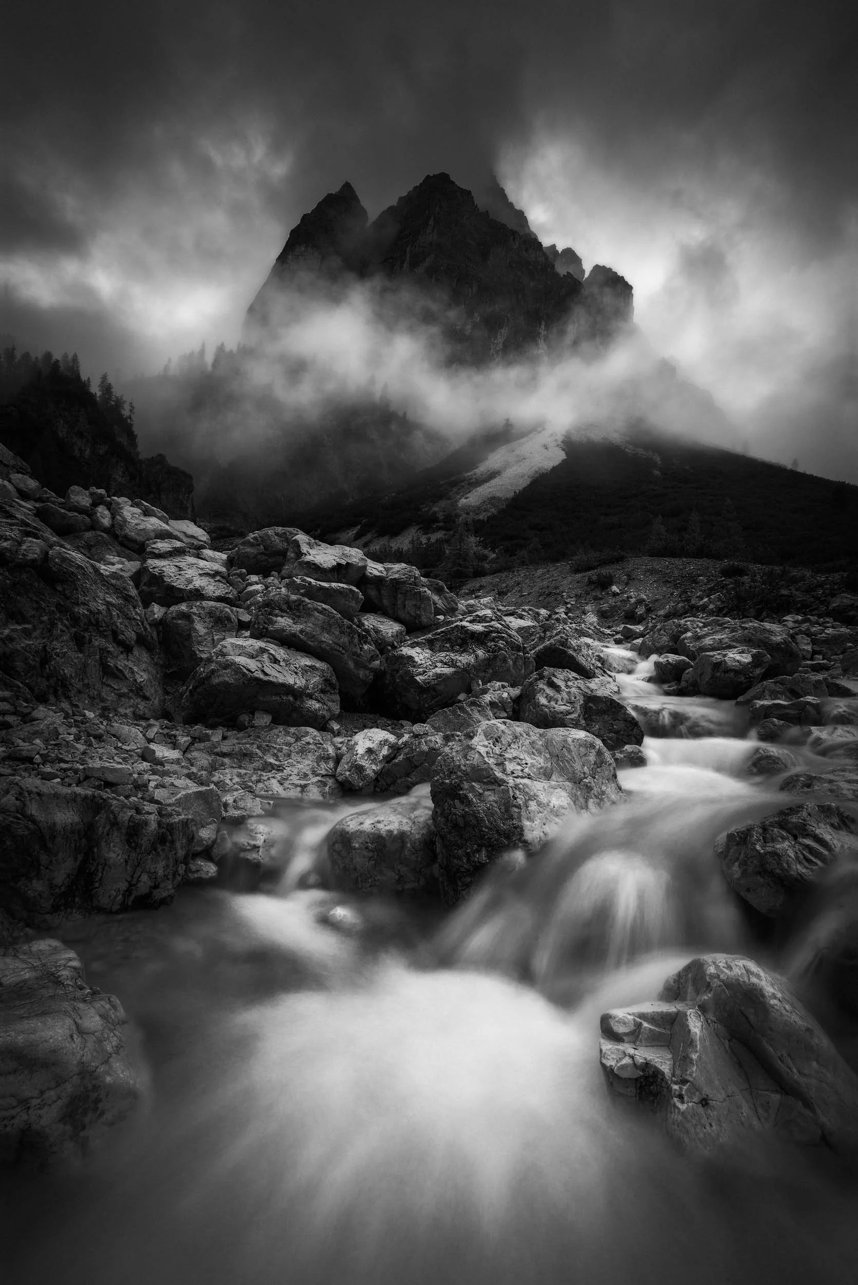 Dramatic black and white image of a river cascade in front of a dark mountain in the Dolomites surrounded by clouds. 