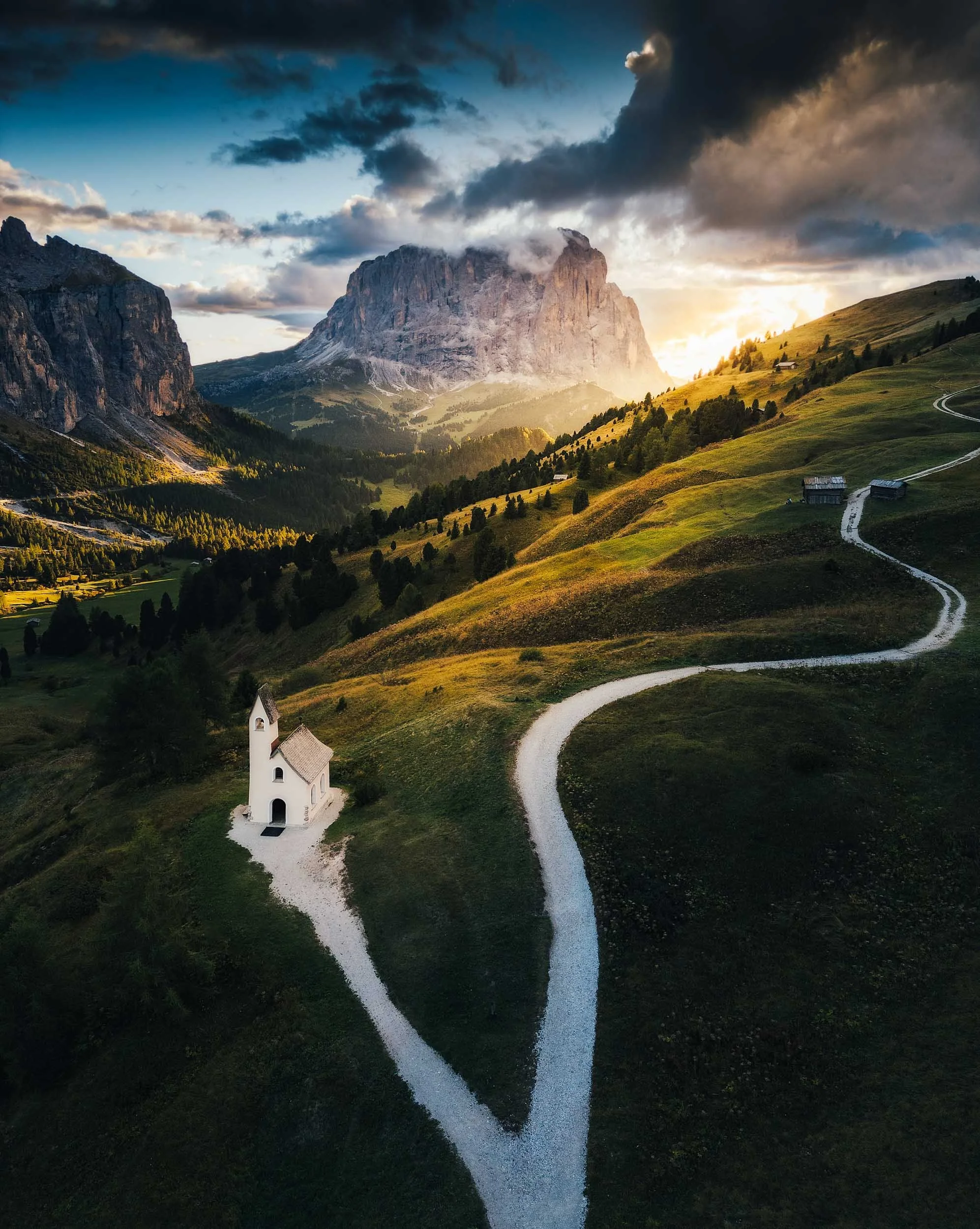 Aerial picture of chapel and winding mountain path at Passo Gardena in the Dolomites with Sassolungo in the background during sunset.
