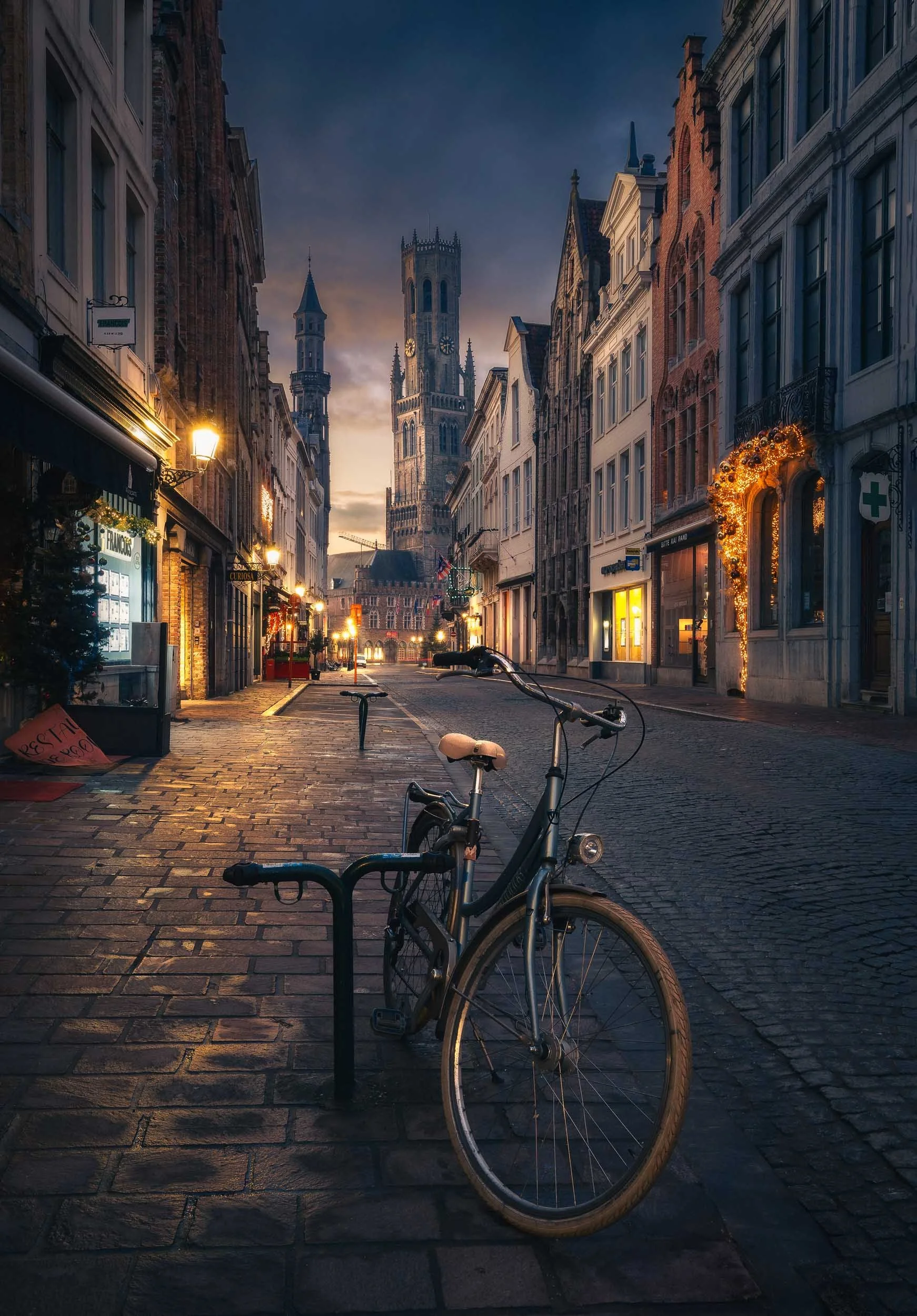Bicycle in the streets of Bruges shortly before sunrise, with the streets illuminated and Belfort towering in the background.