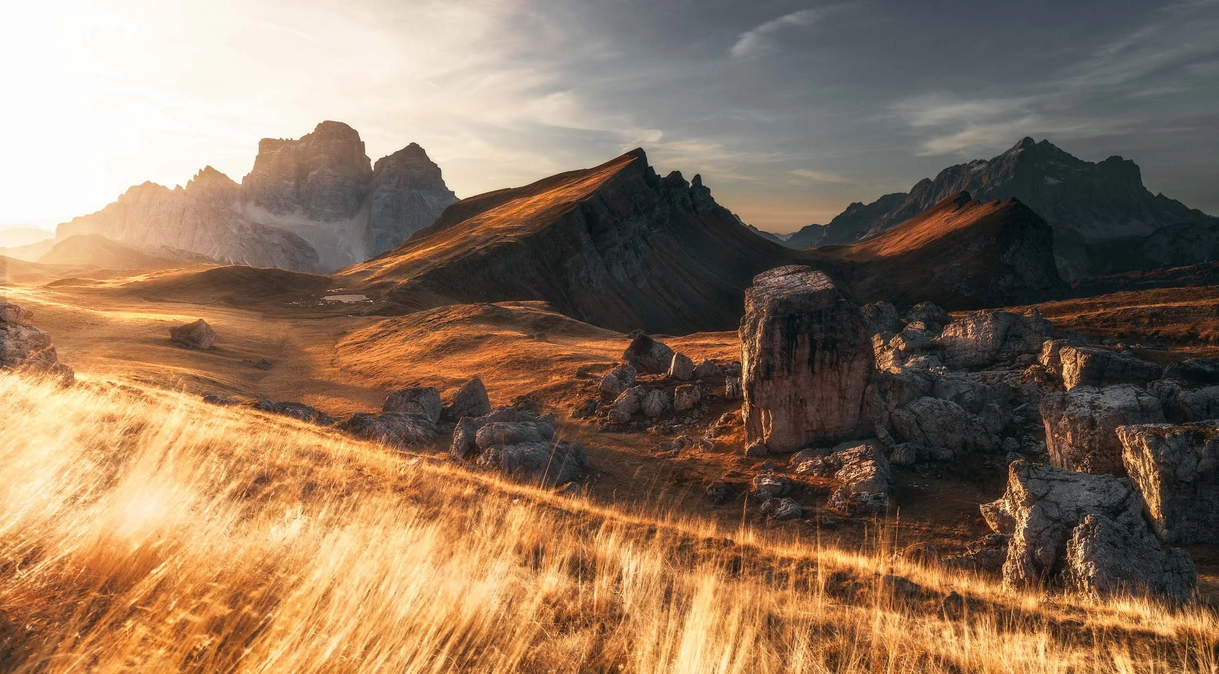 Malga Mondeval with its big boulders scattered in the valley during a warm autumn sunrise with Monte Pelmo towering in the background and the grass in the foreground being lit by the sun. 
