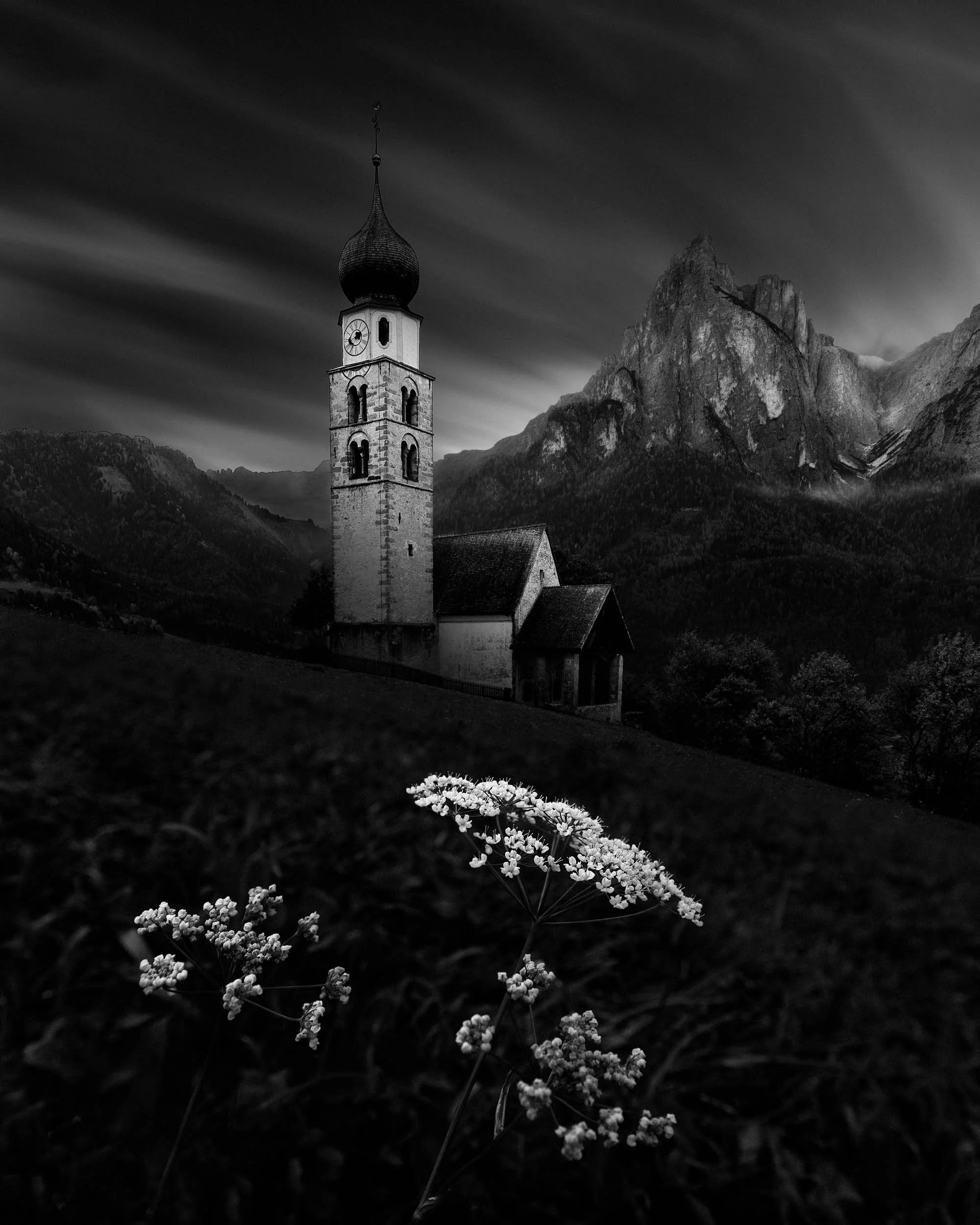Black and white long exposure of St. Valentin church with the Schlern massif in the background and some flowers in the foreground.