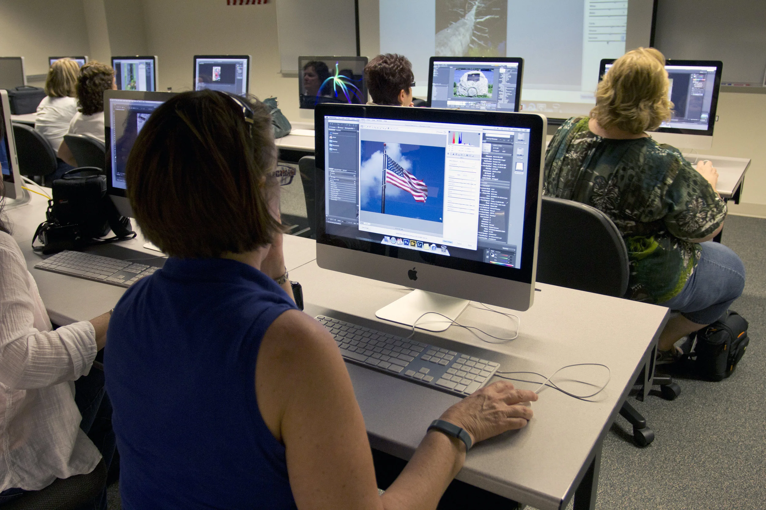 Photograph of a group of people sitting in a classroom setting. Everyone is seated behind a computer. View of the back of their heads as they look forward at an image projected on a screen.