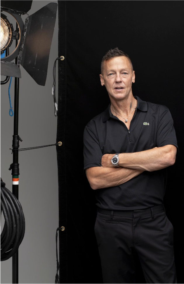 Robert Maxwell standing in a studio space with a studio light next to him. He has his arm crossed with a relaxed facial expression