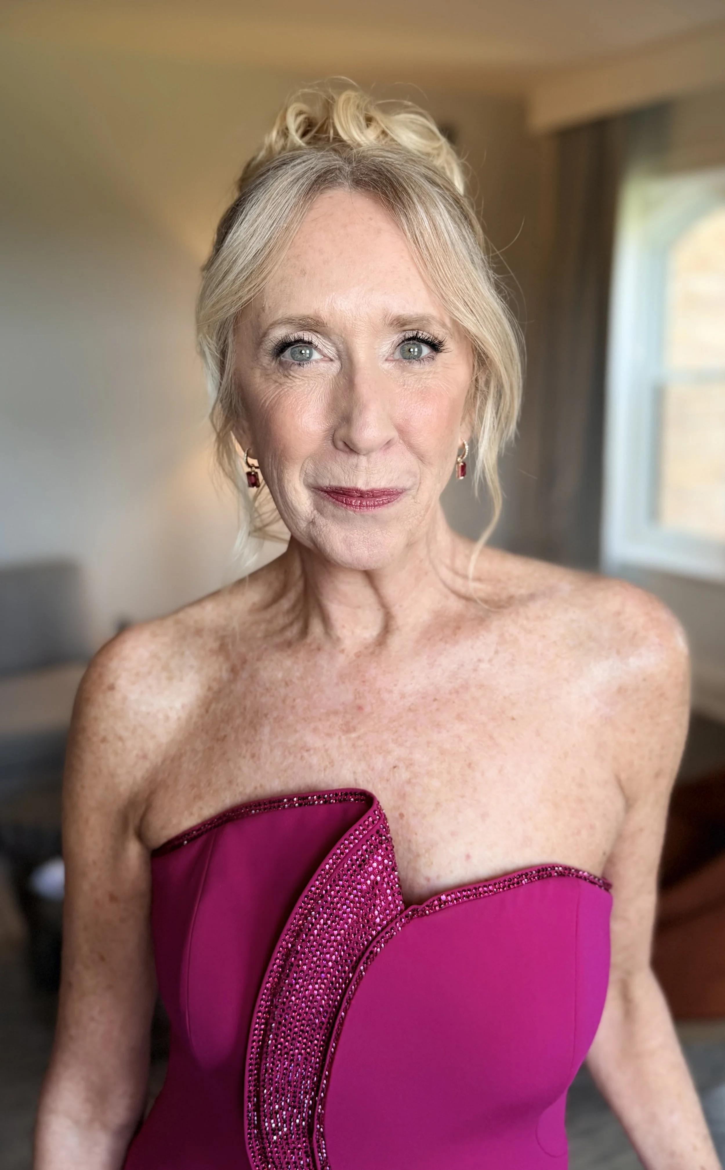 A mature woman with blonde hair styled up wears a strapless magenta dress with rhinestone embellishments, standing near a window in a softly lit room. Mother-of-the-bride hair and makeup by Leather & Lace Artistry.