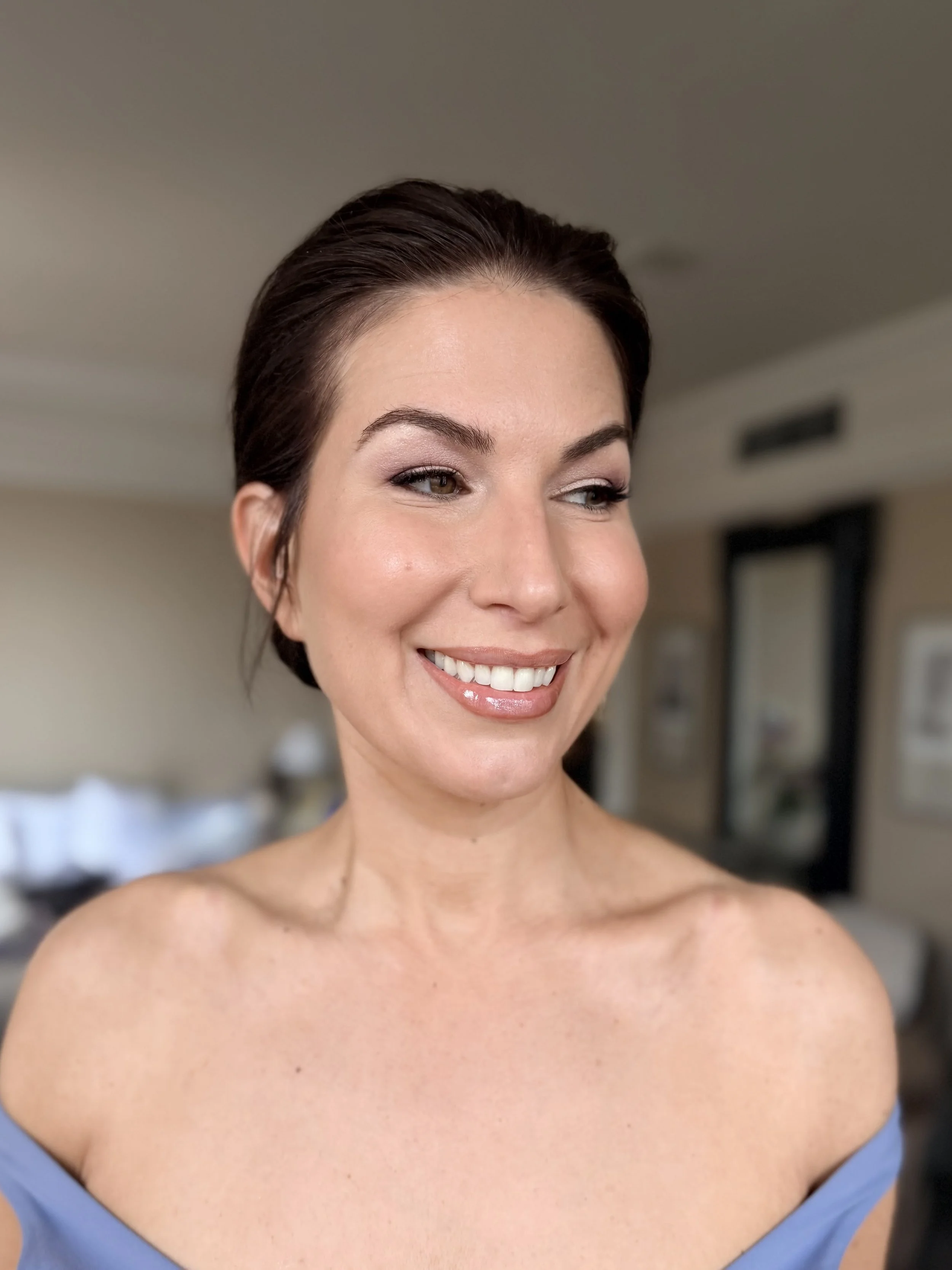 A smiling woman with short dark hair styled away from her face, wearing a blue off-shoulder top, standing indoors with a neutral background. Bridal hair and bridal makeup by Leather & Lace Artistry.