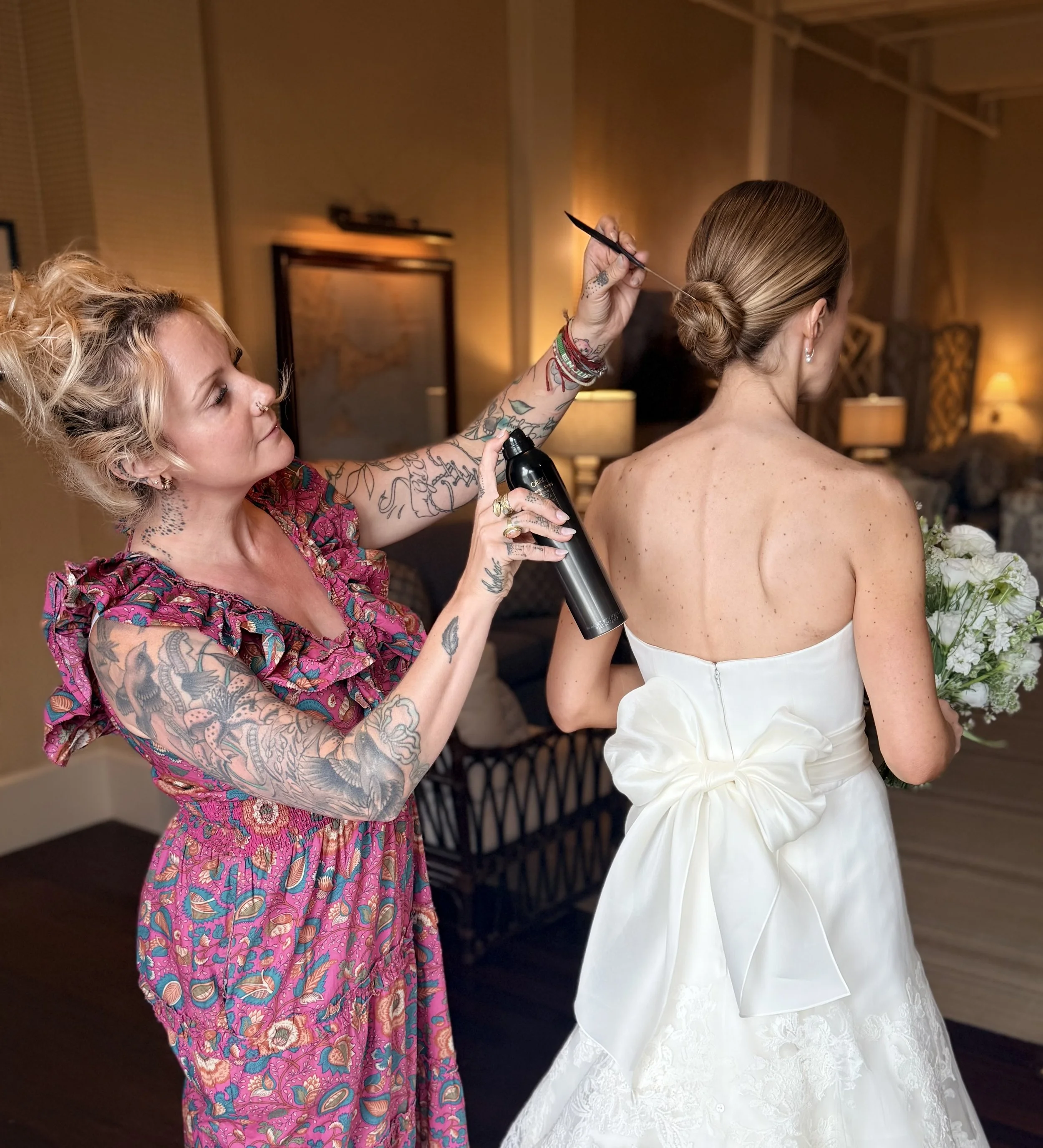 A woman in a sleeveless white wedding dress has her hair styled in an elegant bun and is being prepared for her wedding. Bre Welch of Leather & Lace Artistry is using a spray bottle and holding a hairpin or tool, working on the bride's hair.