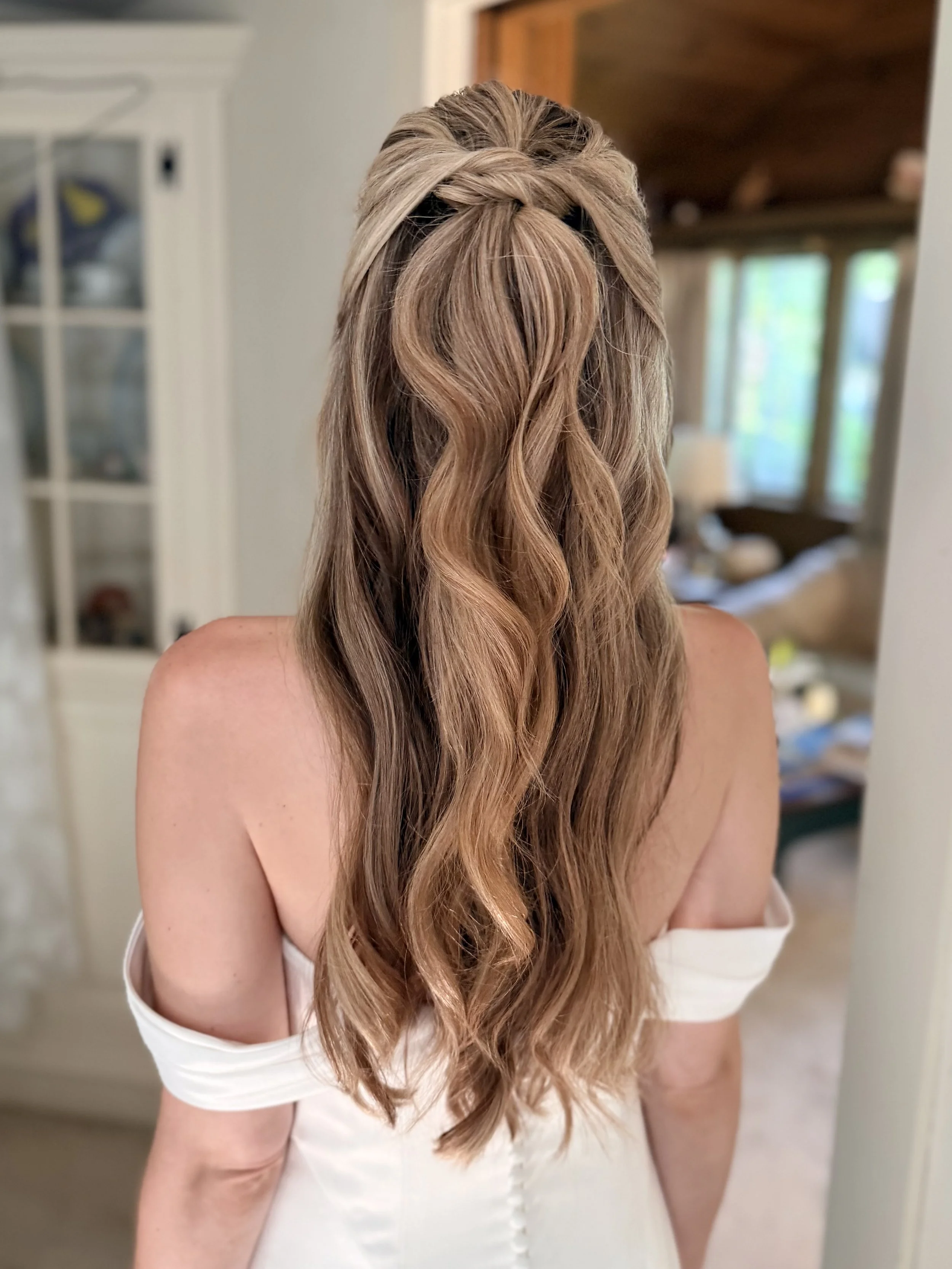 Back view of a woman with long wavy light brown hair styled in a half-up, braided hairstyle, wearing a white off-shoulder dress in a room with natural light, a mirror, and a cozy interior. Leather & Lace Artistry created her bridal hairdo.