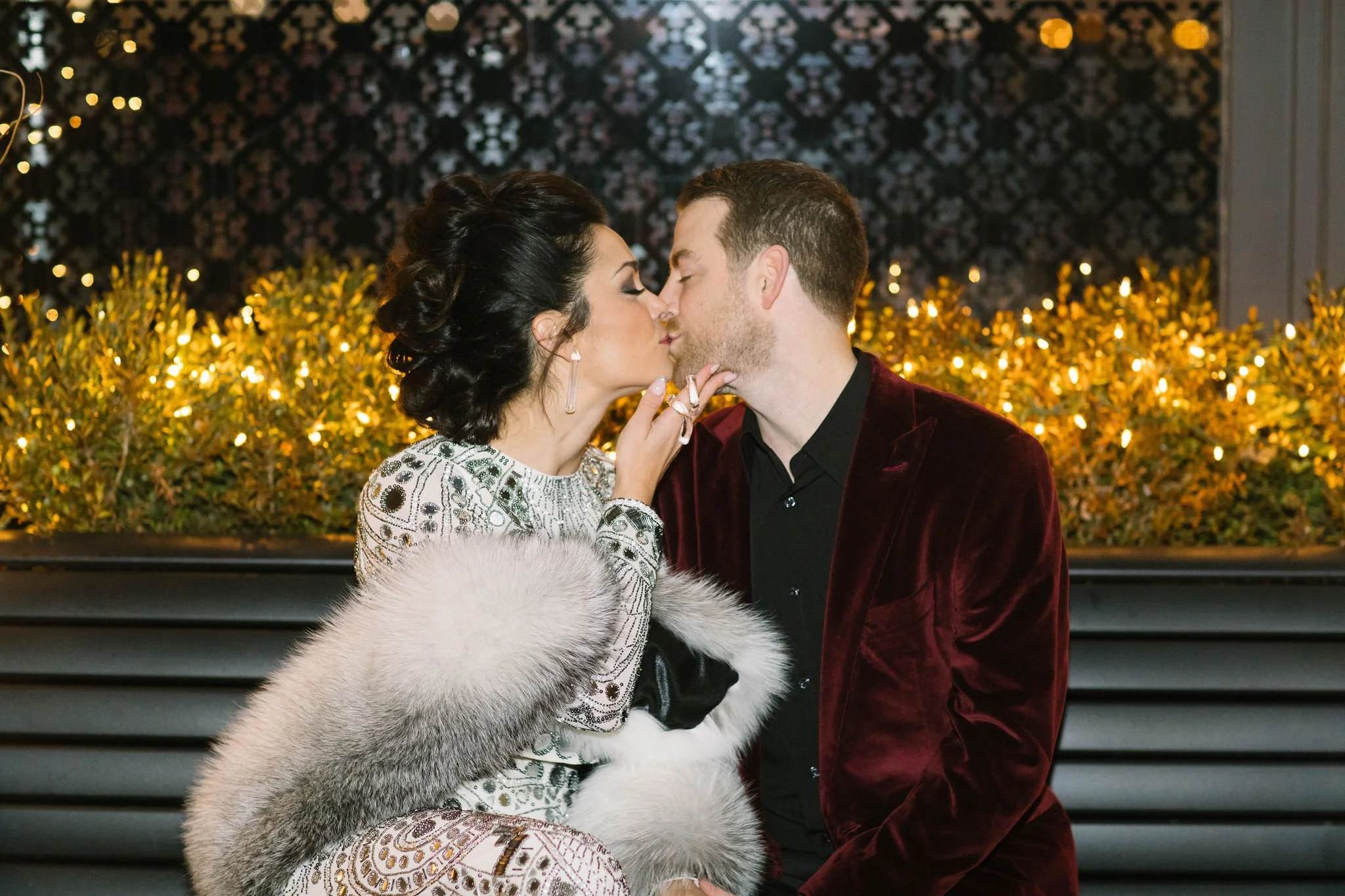 A woman and a man are about to kiss. The woman is wearing a fur shawl and an elegant dress, while the man is dressed in a dark shirt and a velvet blazer. Wedding hair and makeup by Leather & Lace Artistry in New England.