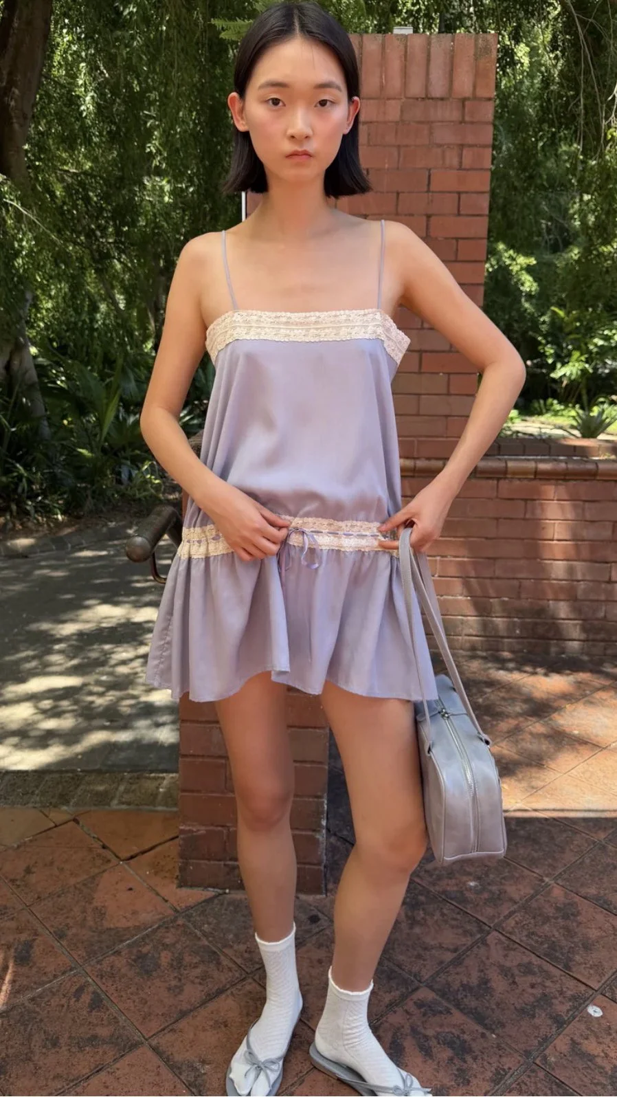 Model wearing a lilac mini dress with cream lace trim, white socks, pale blue bow flats, and a matching handbag beside a brick pillar.