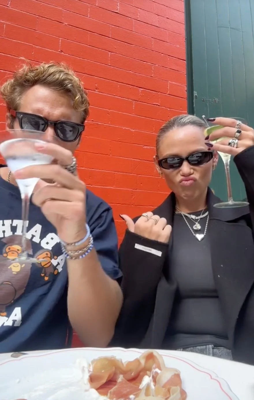 Two people wearing sunglasses hold up cocktails at an outdoor table against a red brick wall, with a plate of food in front of them.