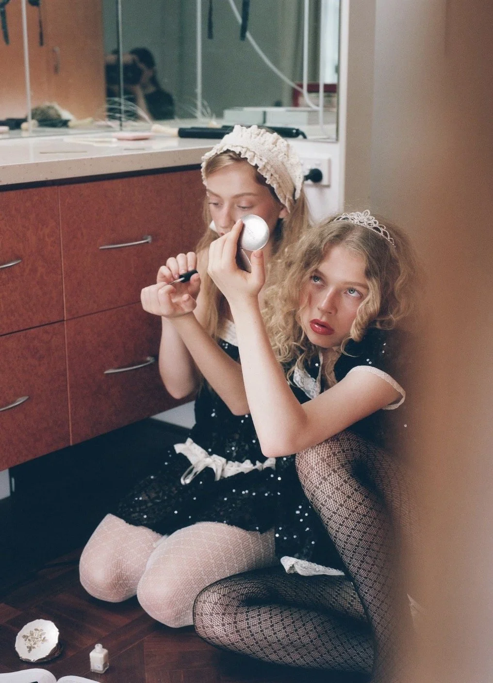 Two models sit beside a bathroom cabinet in black outfits with white lace trim, wearing a ruffled bonnet and a tiara while getting ready.