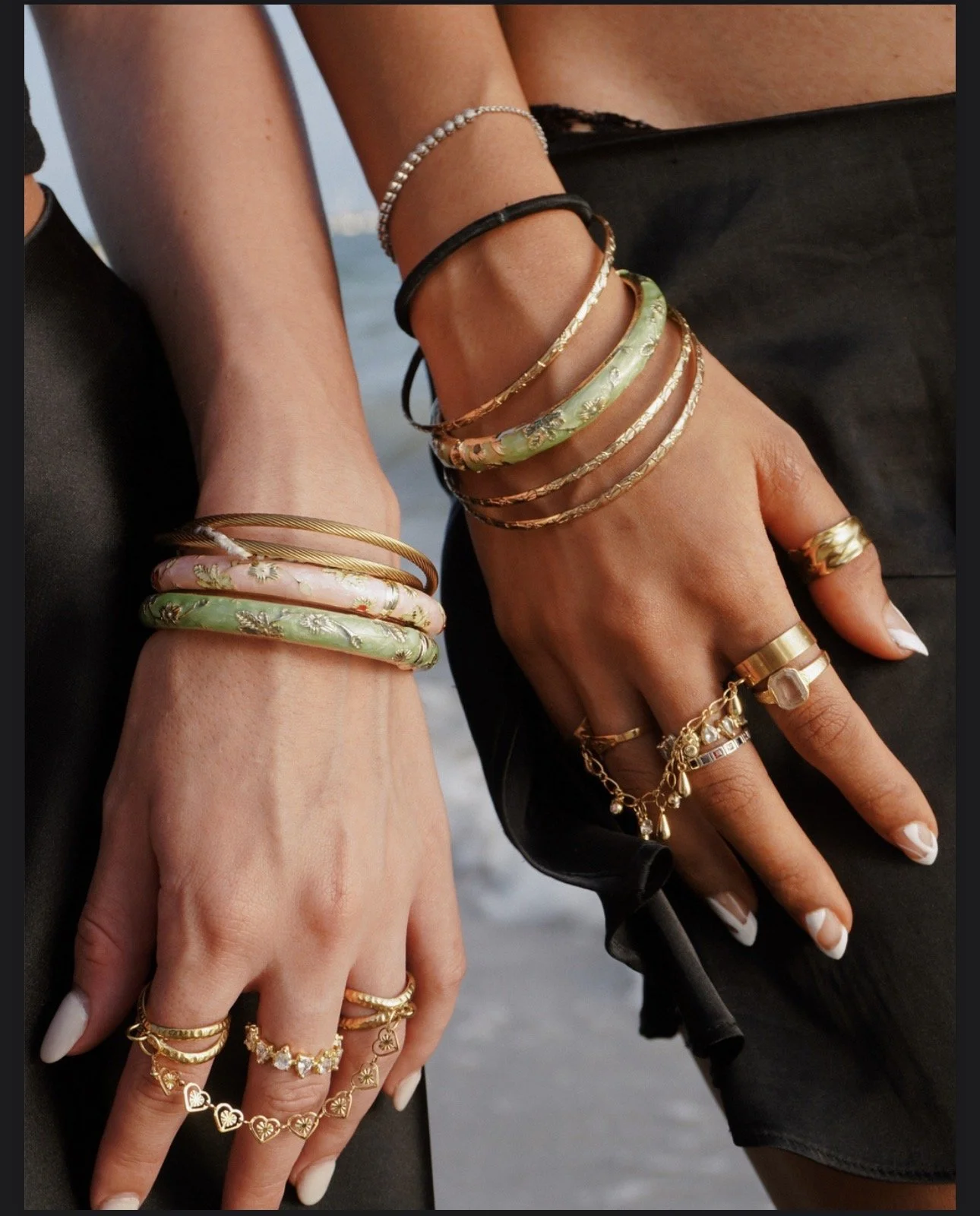 Two women’s hands styled with layered gold rings, hand chains and pastel bangles by Amoura Jewelers.