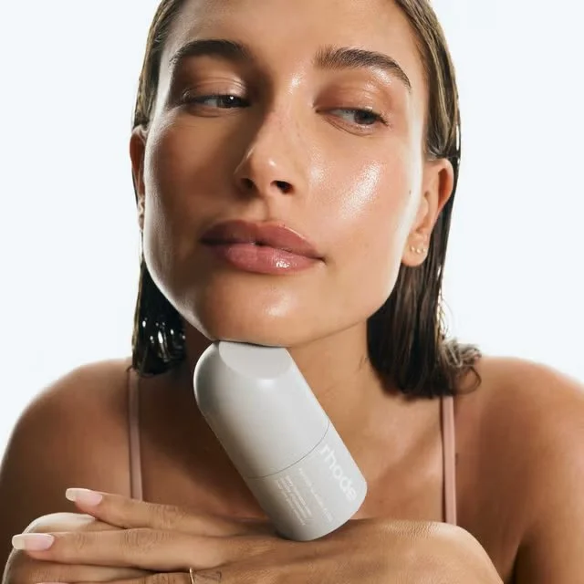 Girl with slicked-back hair and glossy skin rests her chin above her hands, with a pale Rhode skincare bottle beneath her face.