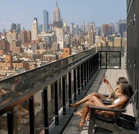 Two women reclining in chairs on a sunny balcony overlooking Manhattan, with the Empire State Building in the distance.