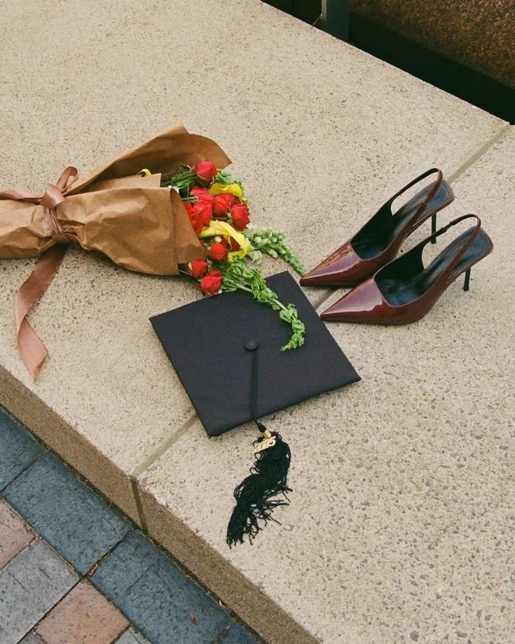 A black graduation cap beside a bouquet of flowers and burgundy slingback heels on stone steps.