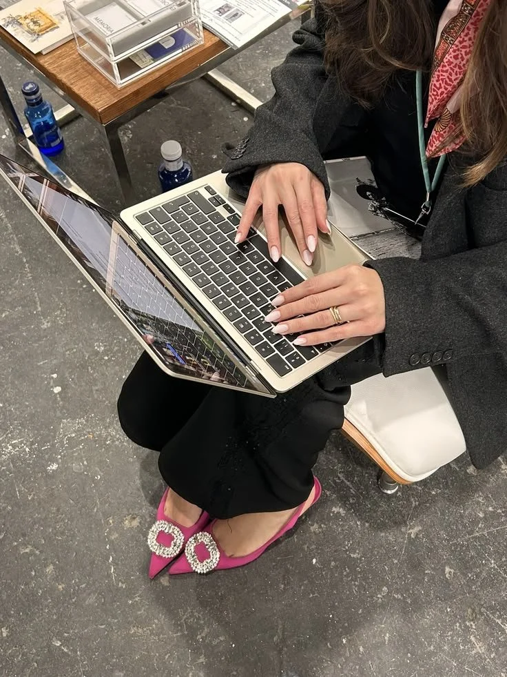 A woman typing on a laptop while wearing a black suit and bright pink jeweled heels.