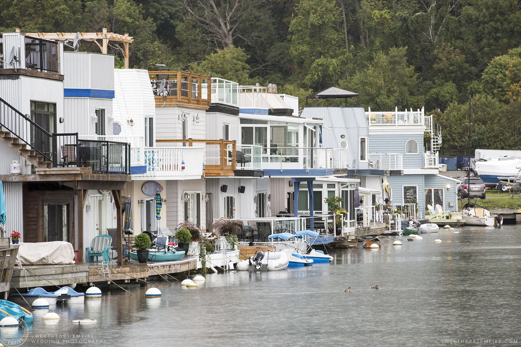 A row of houseboats in nautical-colours, at the Bluffers Park Marina, with the greenery of Scarborough Bluffs rising off in the background, on a rainy day.