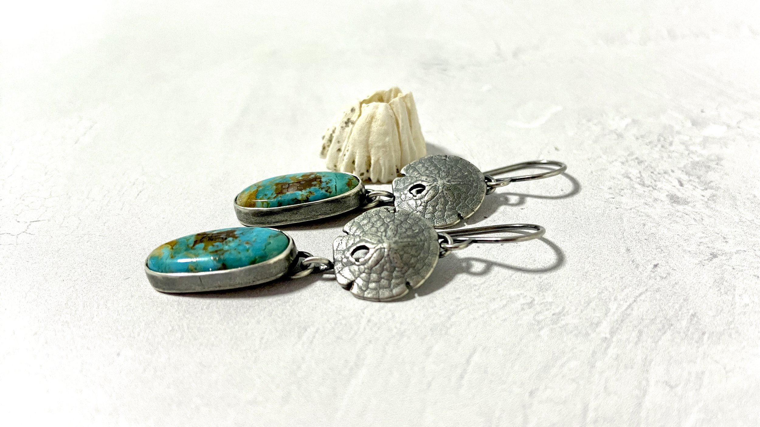 A pair of sand dollar earrings with Mohave Turquoise oval drops are displayed elegantly alongside a real barnacle