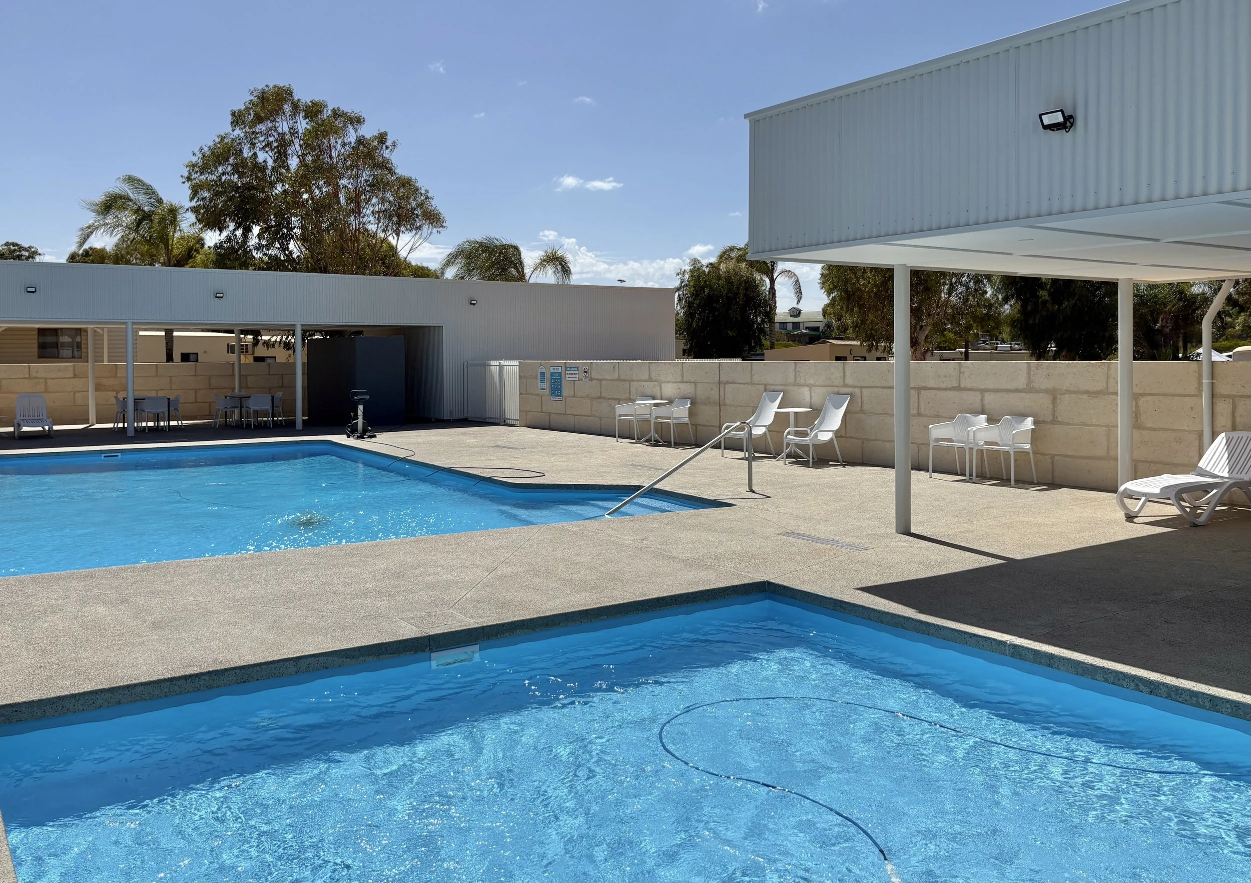 Empty outdoor swimming pool with a smaller hot tub in the foreground, poolside chairs and tables, covered shaded area, and a beige wall with trees behind it under a clear blue sky.