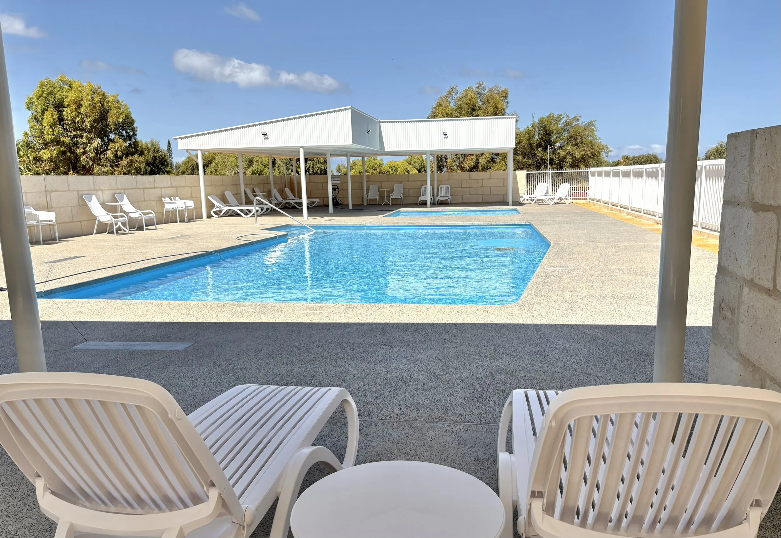 Empty outdoor swimming pool with lounge chairs, a shaded area, and a white fence under a blue sky with some clouds.