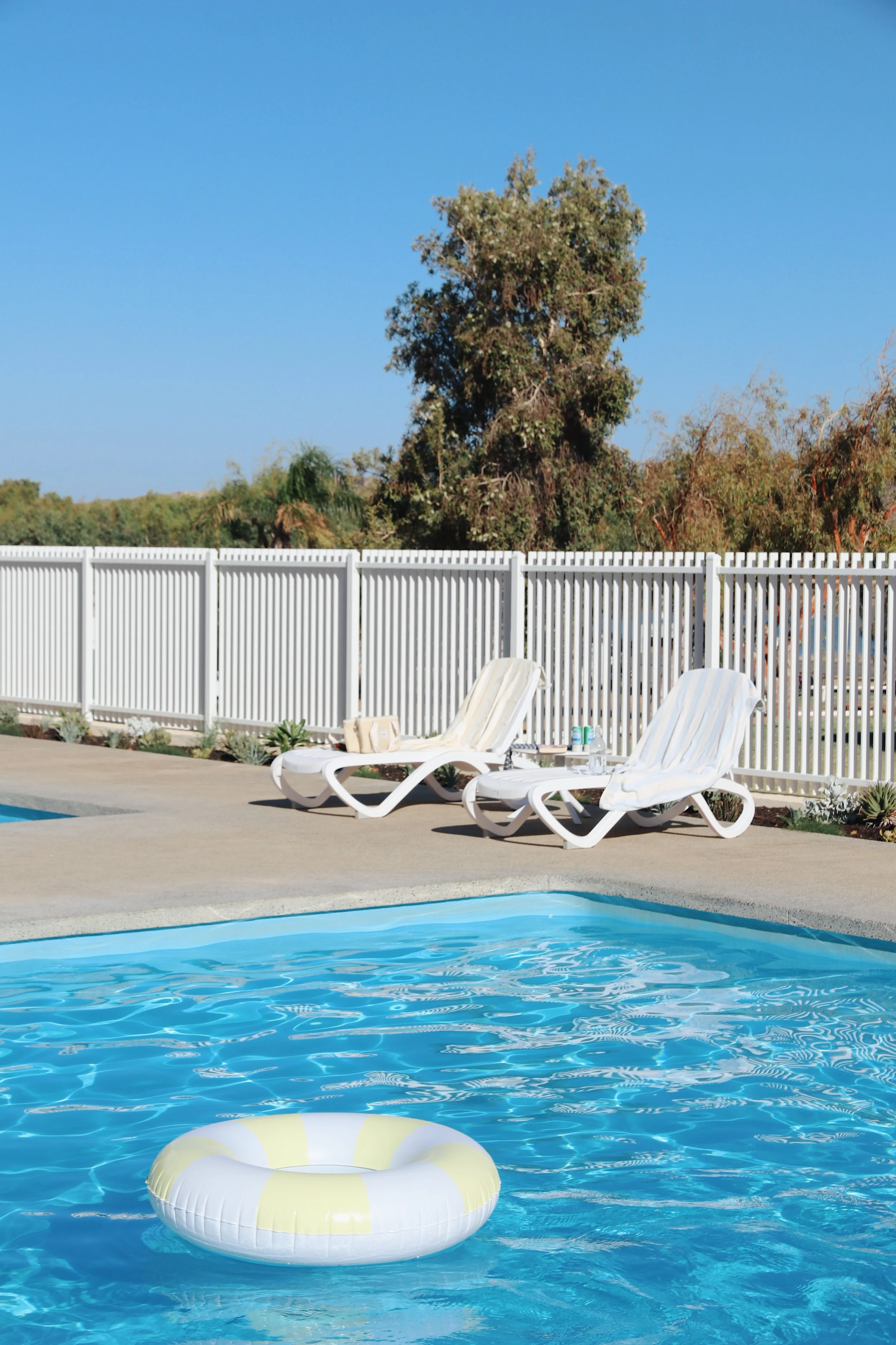 Two white lounge chairs with striped cushions beside a swimming pool with a yellow and white inflatable ring. A white fence surrounds the pool area, with a large tree and blue sky in the background.