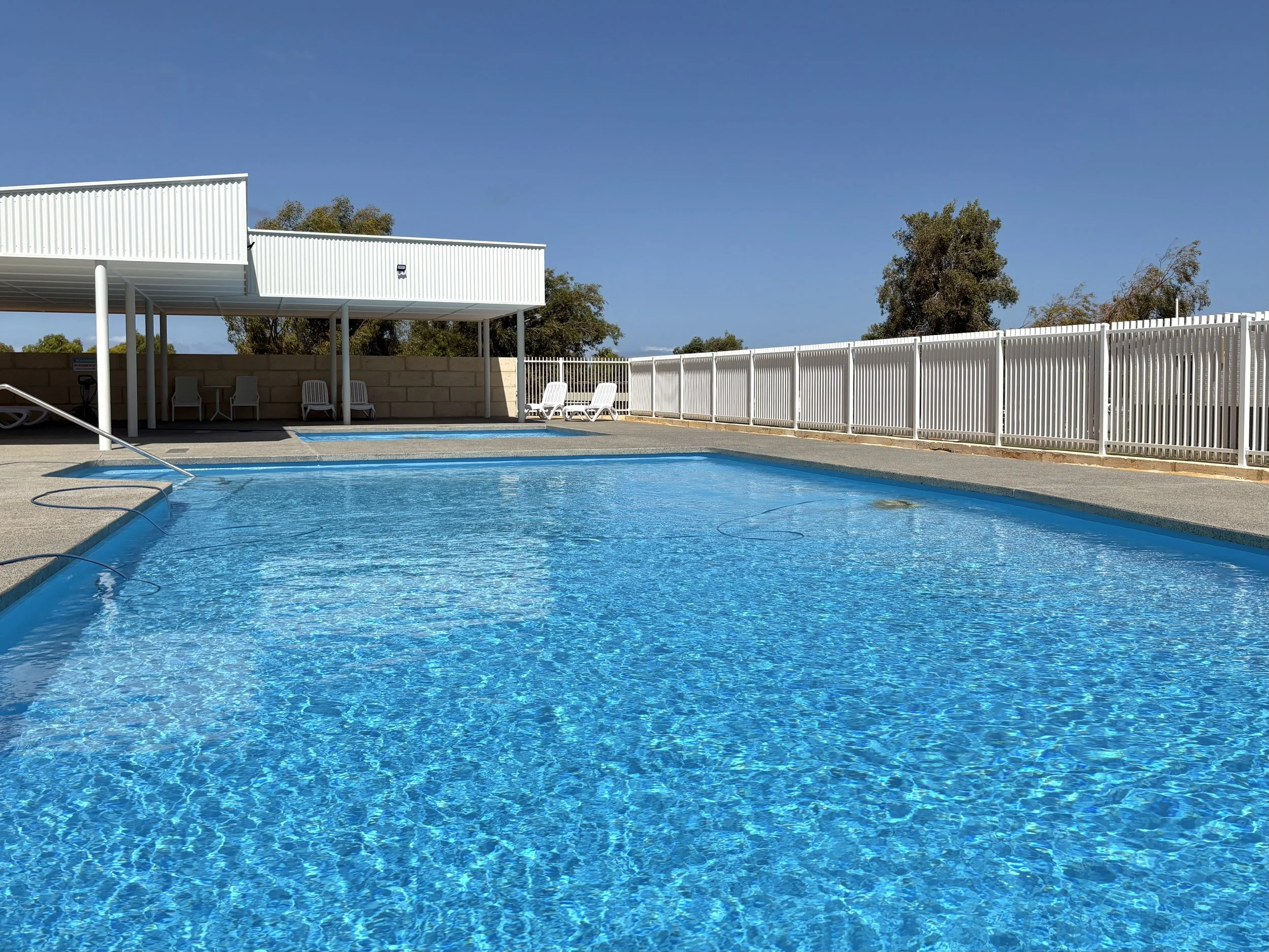 Empty outdoor swimming pool with blue water, surrounded by white fence and lounge chairs, under a clear blue sky.