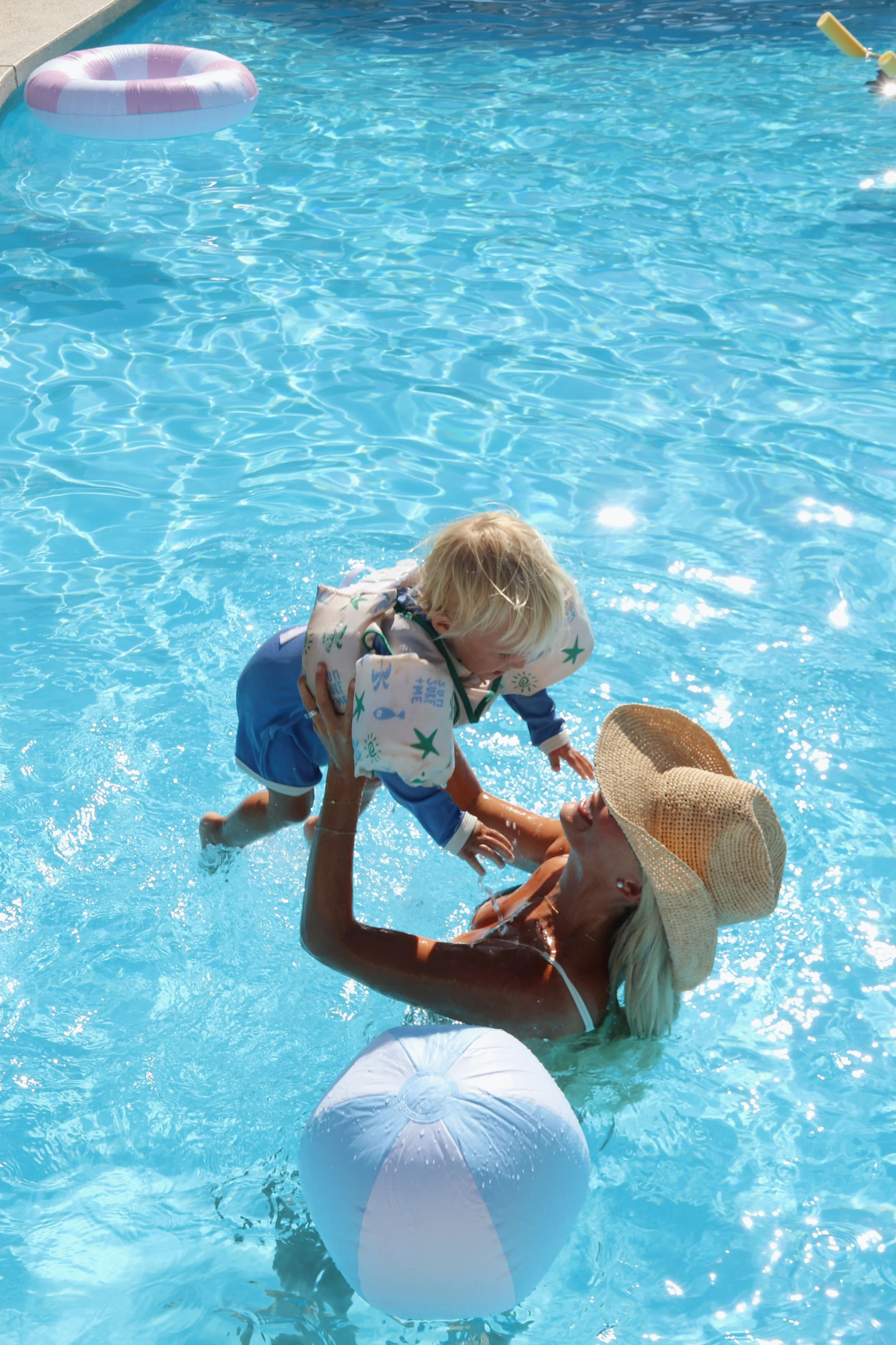 A woman and a young child playing in a swimming pool, with the woman in the water wearing a large sun hat and the child in a flotation device, surrounded by pool toys and a bright blue pool.