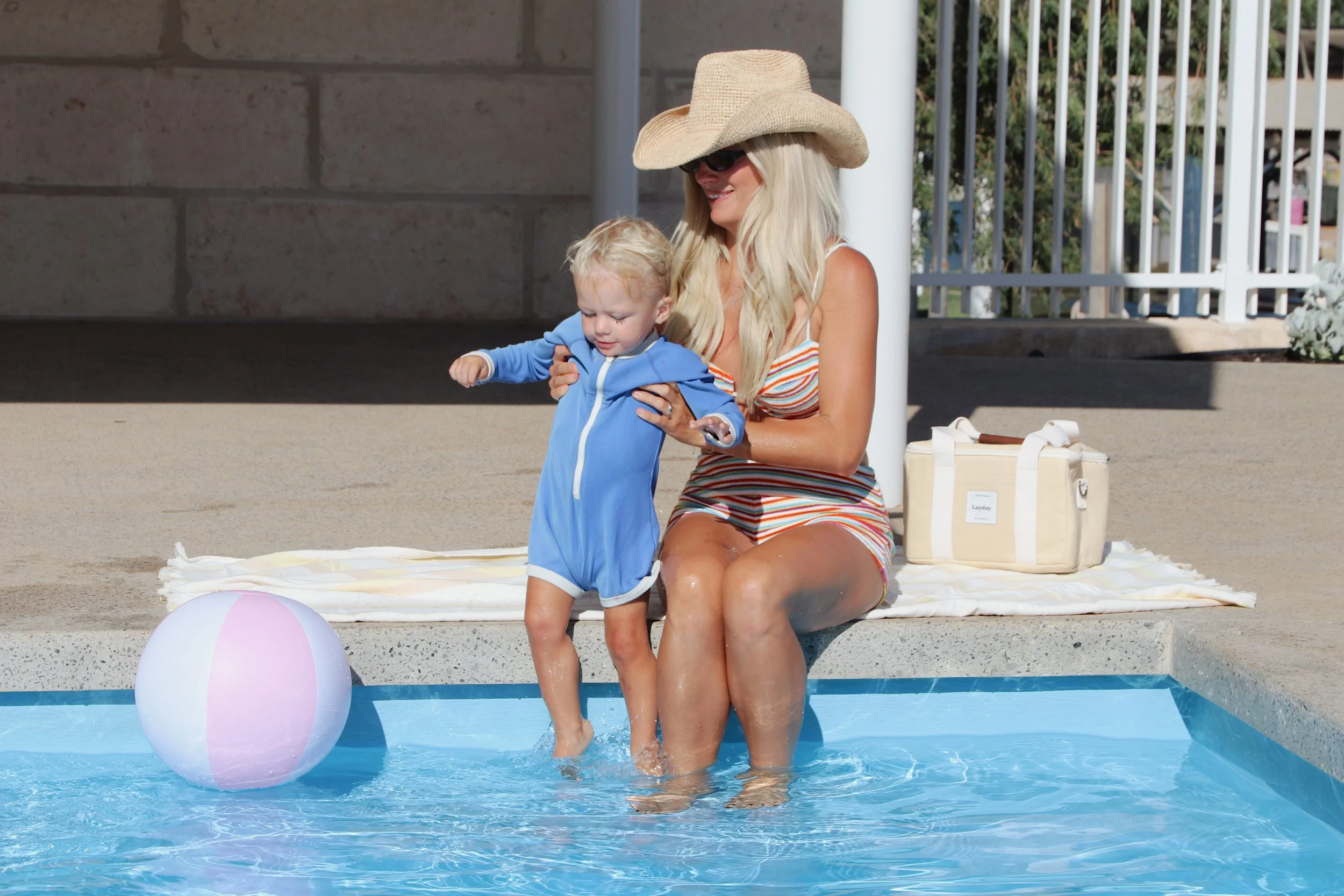 A woman wearing a large straw hat and sunglasses sitting by a swimming pool, holding a young child in a blue swimsuit as they go into the water, with a beach ball near the edge of the pool.