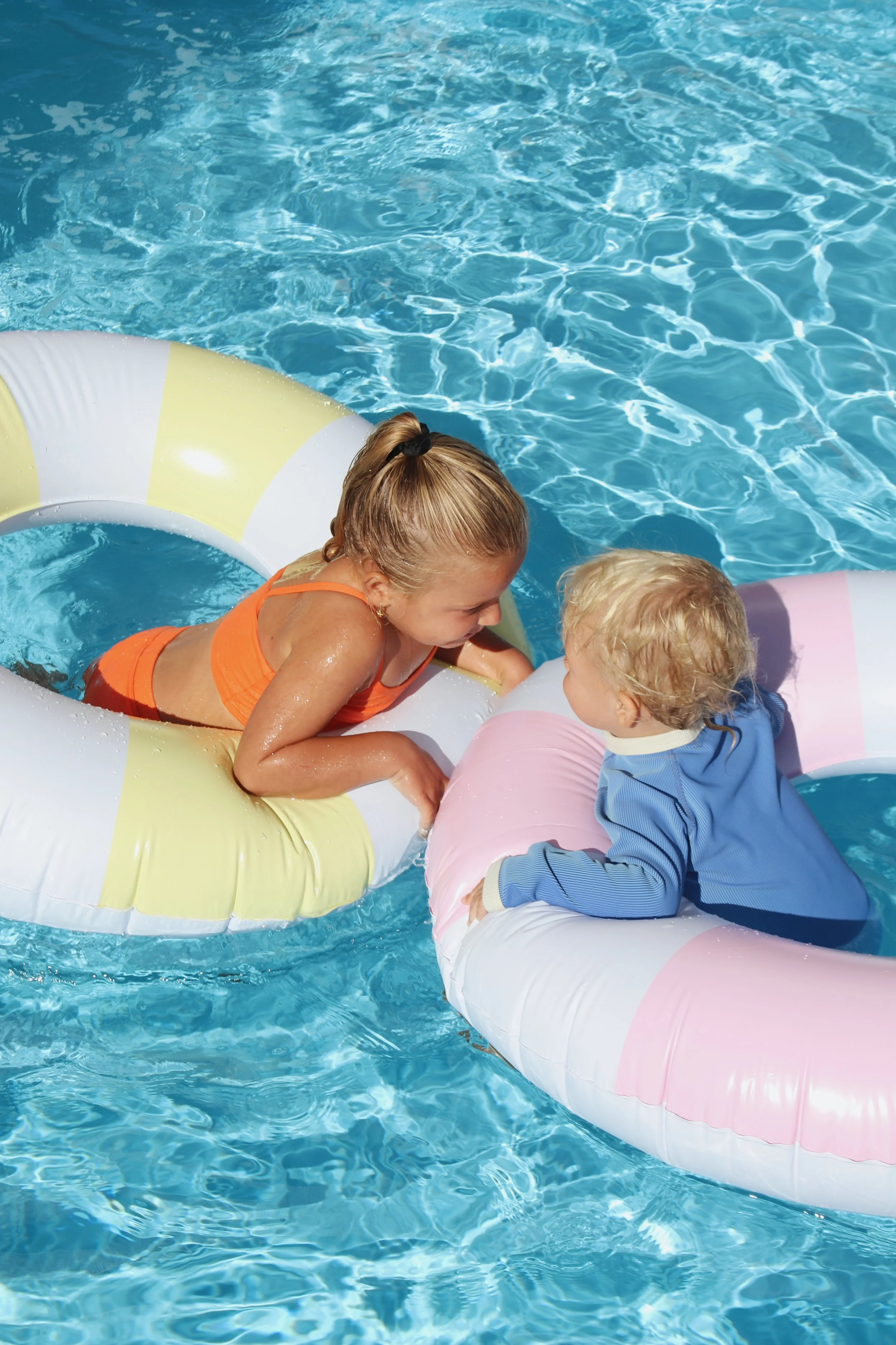 Two young children, a girl and a boy, are playing in a swimming pool on inflatable floats and talking to each other.
