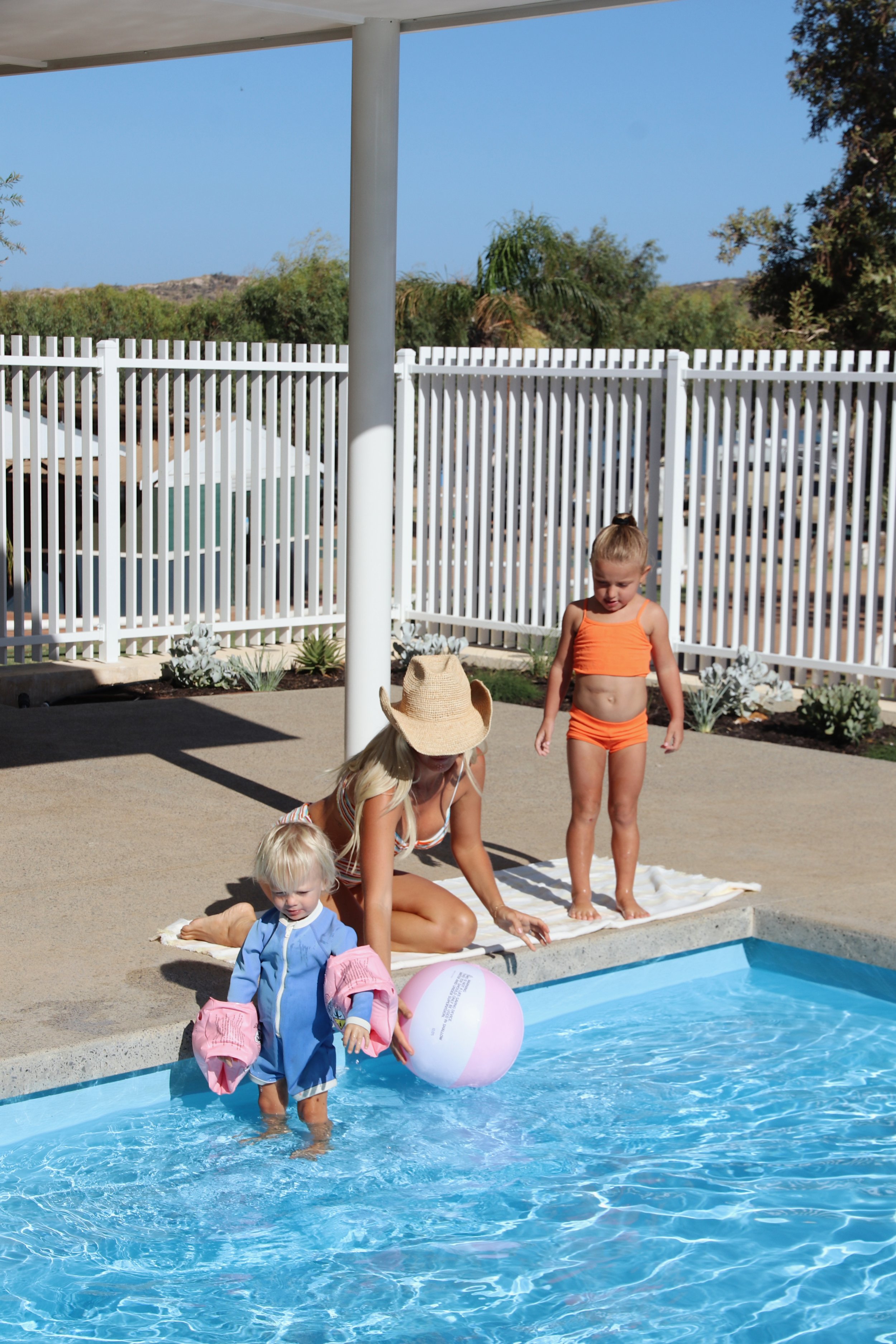 A woman and two young girls at a swimming pool, with the woman helping a toddler into the water, which has a beach ball, and the girls wearing swimsuits and floaties.