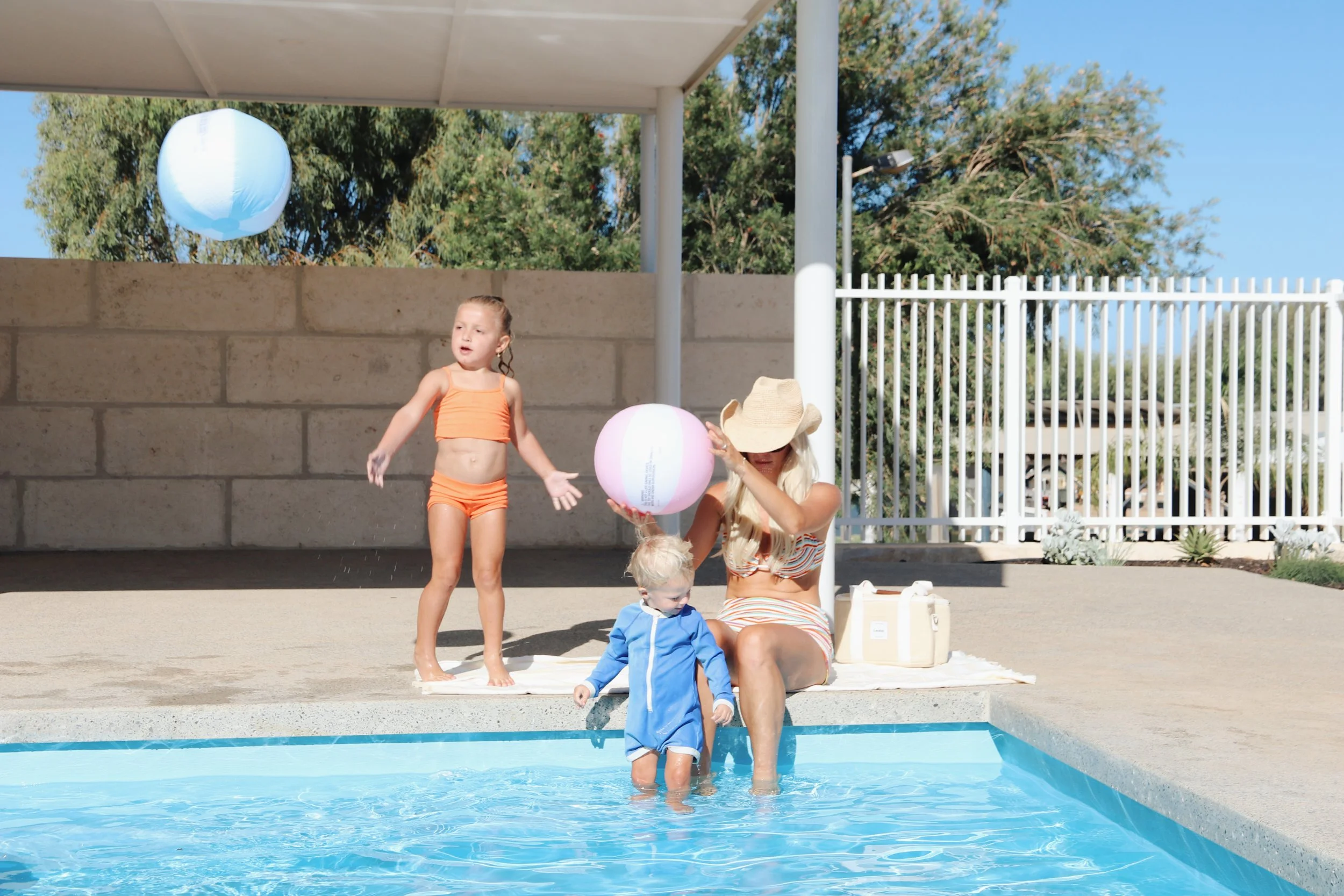 A woman sitting poolside in a striped swimsuit with two children, one sitting on her lap and the other standing nearby, all with beach toys and looking relaxed on a sunny day.