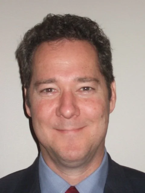 Close-up of a smiling man with dark curly hair wearing a suit and tie.