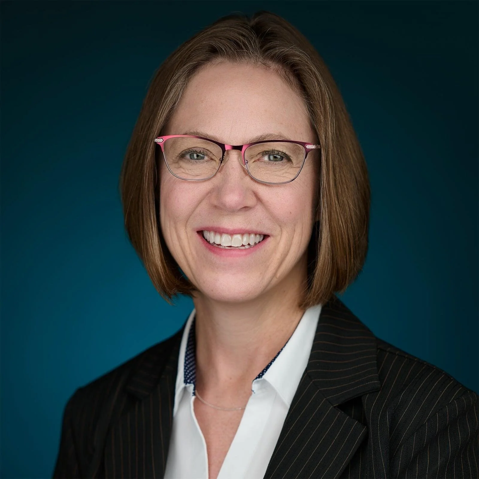 A woman with shoulder-length brown hair, wearing glasses with pink frames, smiling, dressed in a black pinstripe blazer and white shirt, against a blue background.
