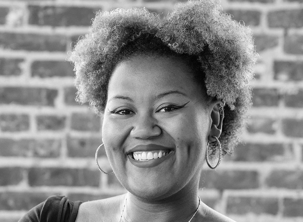 A Black woman with curly hair smiling, wearing hoop earrings and a necklace, standing in front of a brick wall.