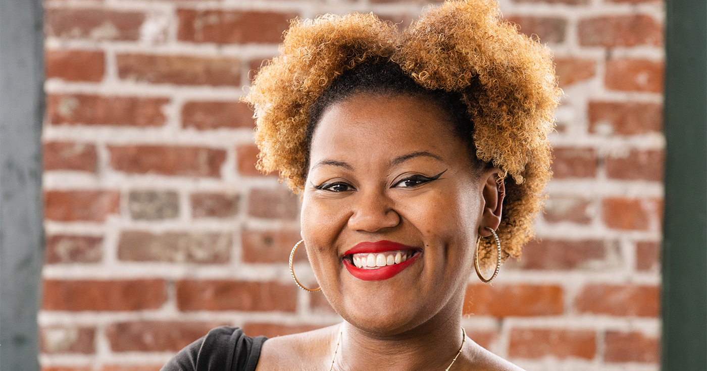 A Black woman with curly hair smiling, wearing hoop earrings and a necklace, standing in front of a brick wall.