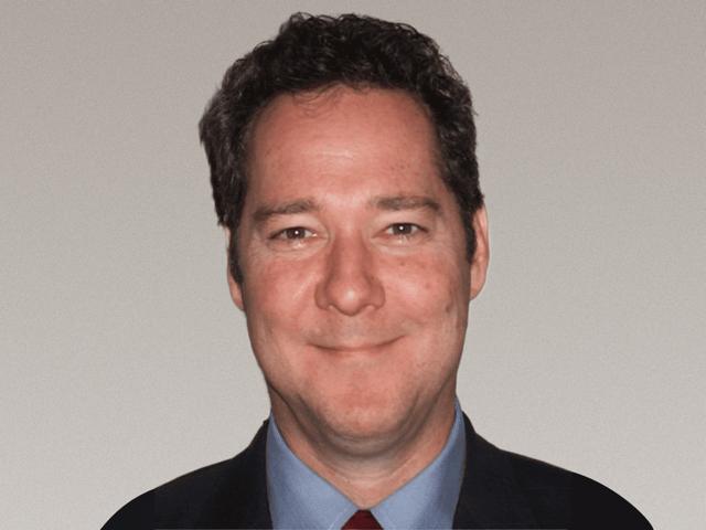 Close-up of a smiling man with dark curly hair wearing a suit and tie.