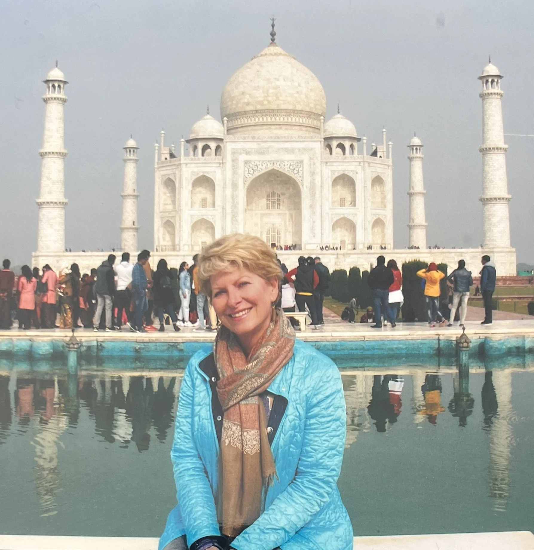 A smiling woman with short blonde hair, wearing a turquoise jacket and a patterned scarf, standing in front of the Taj Mahal in India during daytime, with many tourists and a reflection of the monument in a pool of water.