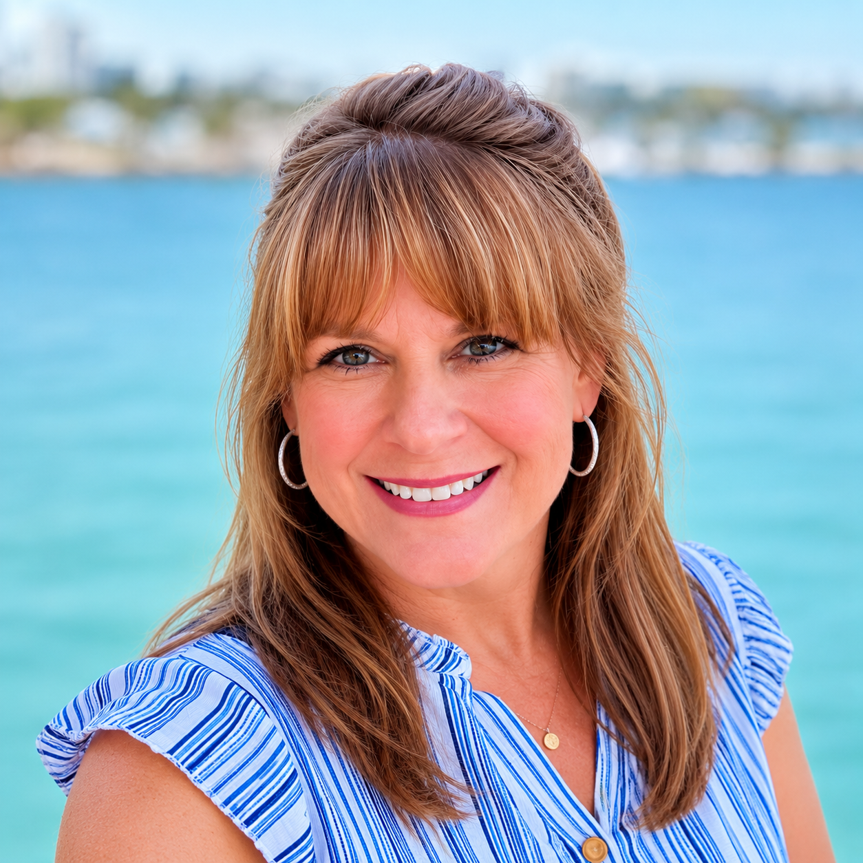 A smiling woman with light brown hair, wearing hoop earrings and a blue and white striped blouse, standing near a body of water with a blurred shoreline in the background.