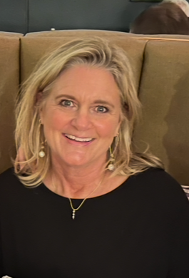 A woman with shoulder-length blonde hair smiling at the camera, wearing a black top, with a beige background and a greenish-blue seating area behind her.