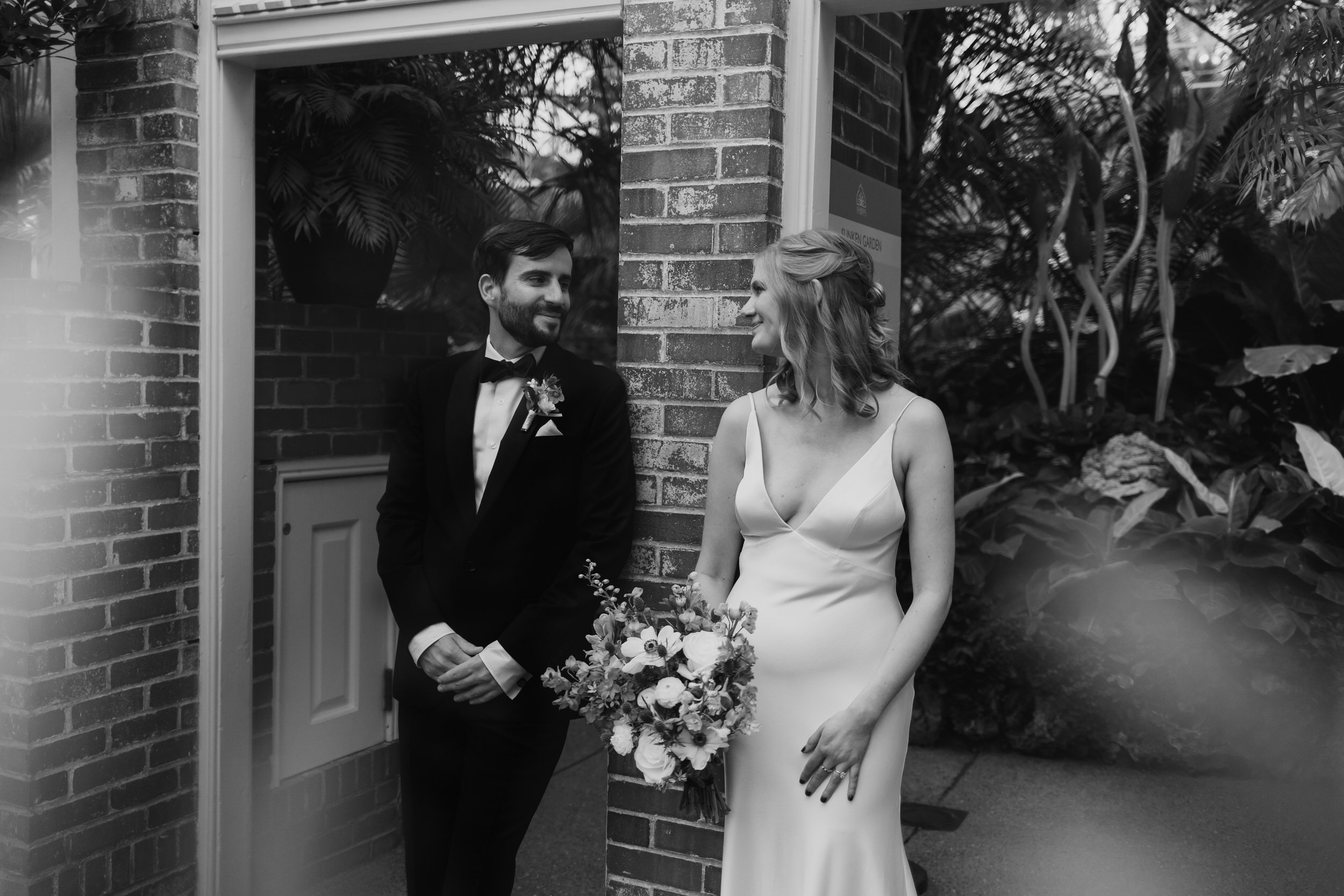 Bride and groom lean against brick wall at Phipps Conservatory while looking at one another.