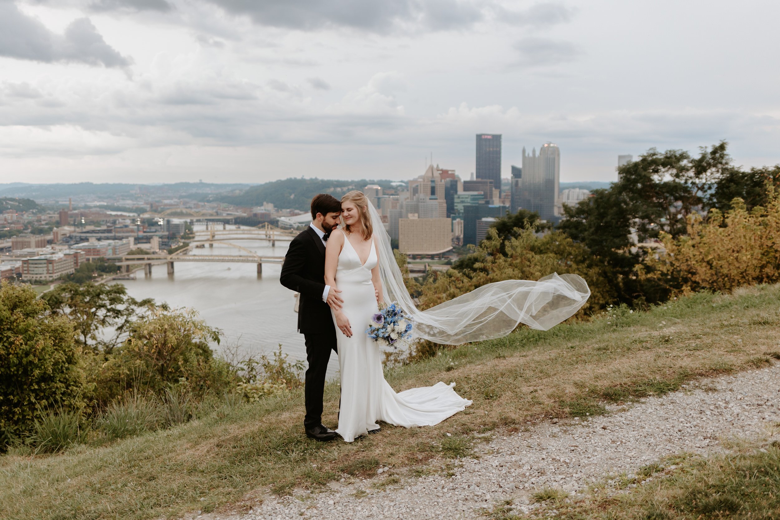 A bride and groom embrace while her veil blows in the wind, with Pittsburgh skyline behind them.