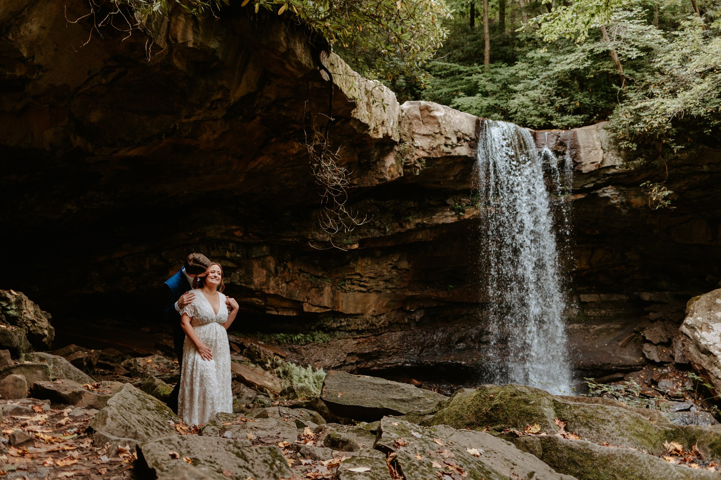 Groom kisses brides cheek while standing in front a waterfall at Ohiopyle State Park.