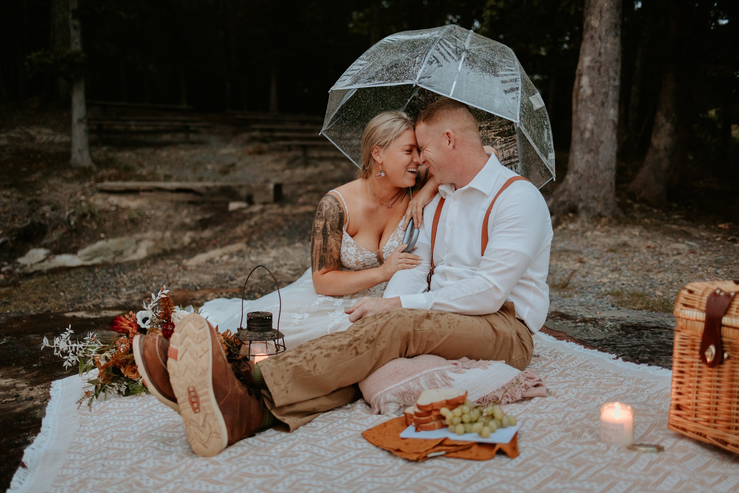 Bride and groom sit on blanket and enjoy candle lit picnic.