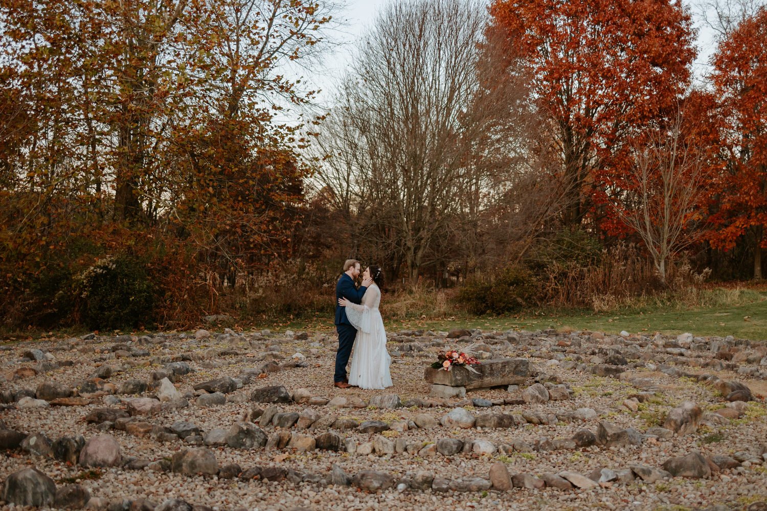 Groom cradles brides face with hands while standing in front of fall trees.
