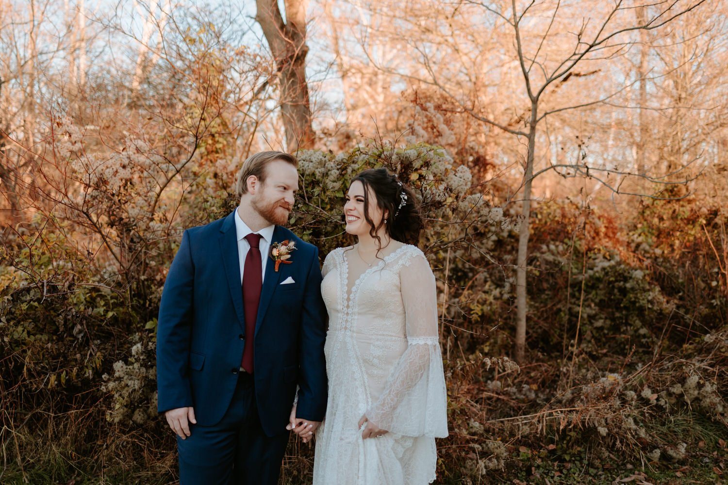 Bride and groom hold hands while smiling and standing in front of fall colored trees.